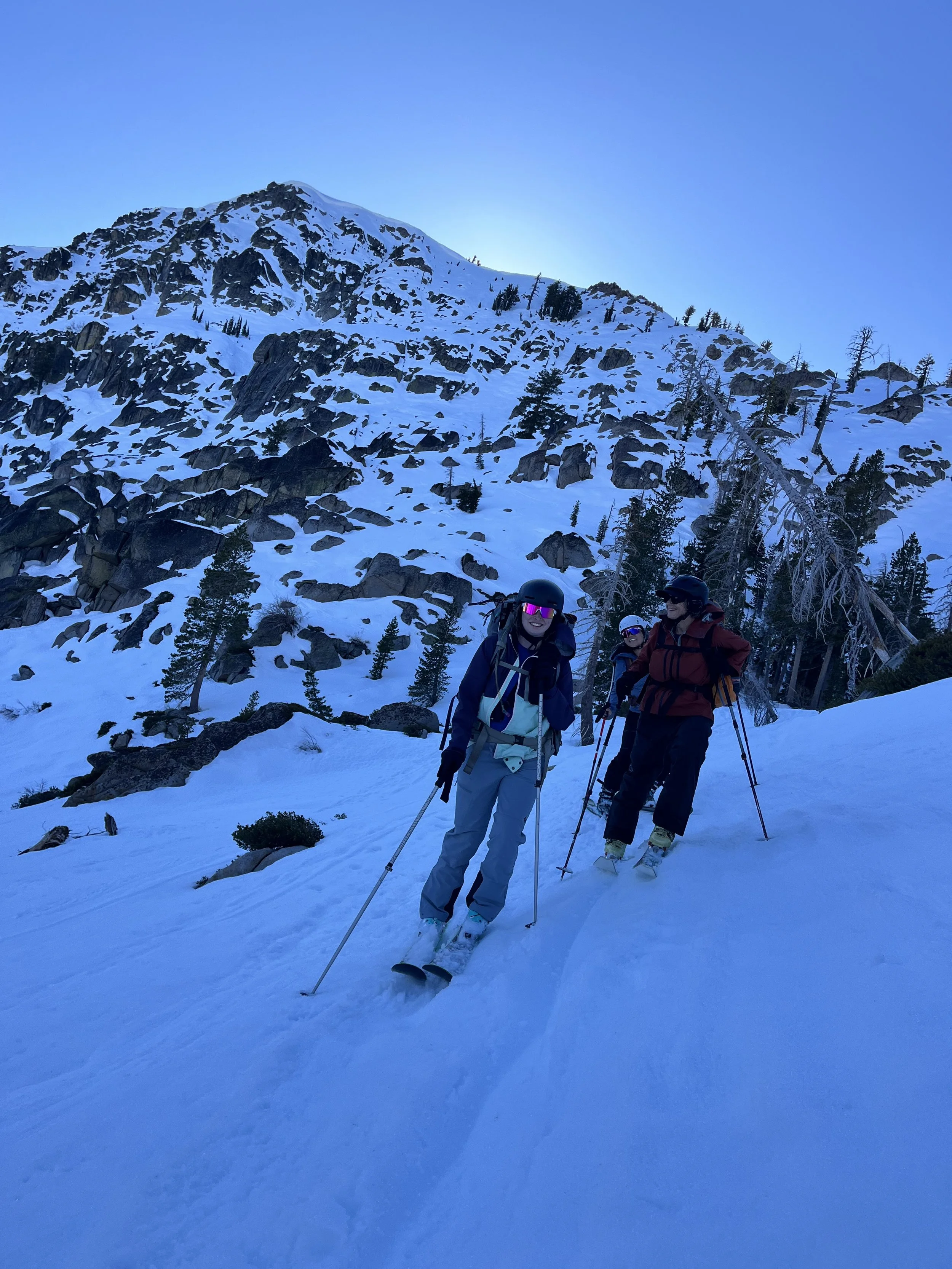 Group of four people in snow-covered mountains, wearing winter gear and ski helmets, standing with ski poles, with rocky mountain and sparse trees in background.