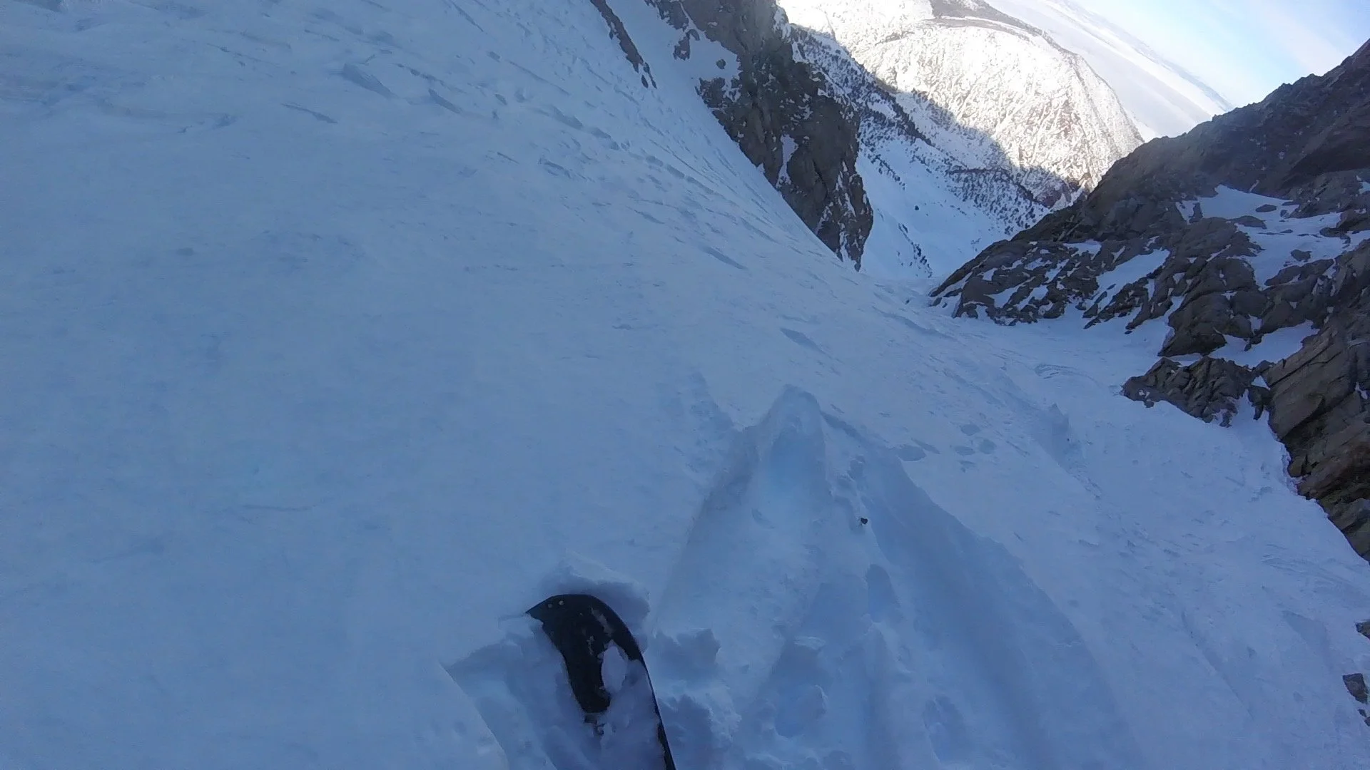 View from a snow-covered mountain slope showing a pair of skis and ski poles in the foreground, rocky cliffs on the right, and tall, snow-capped mountains in the background under a partly cloudy sky.
