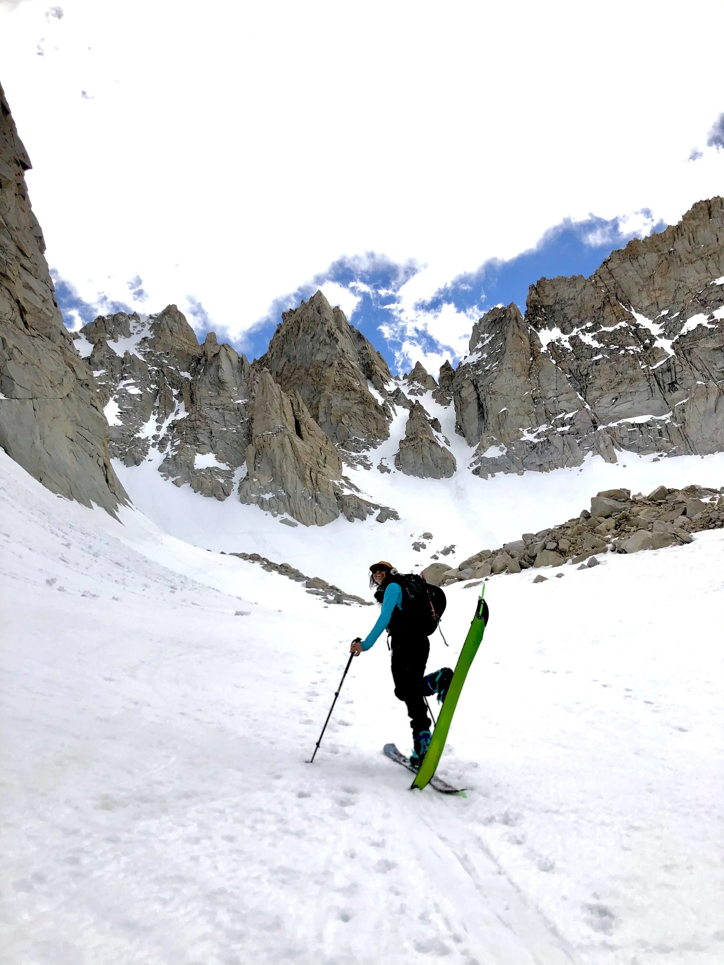 A person in winter gear stands on snow-covered terrain, holding ski poles with a pair of skis attached to their backpack, surrounded by tall rocky mountains and a blue sky with some clouds.