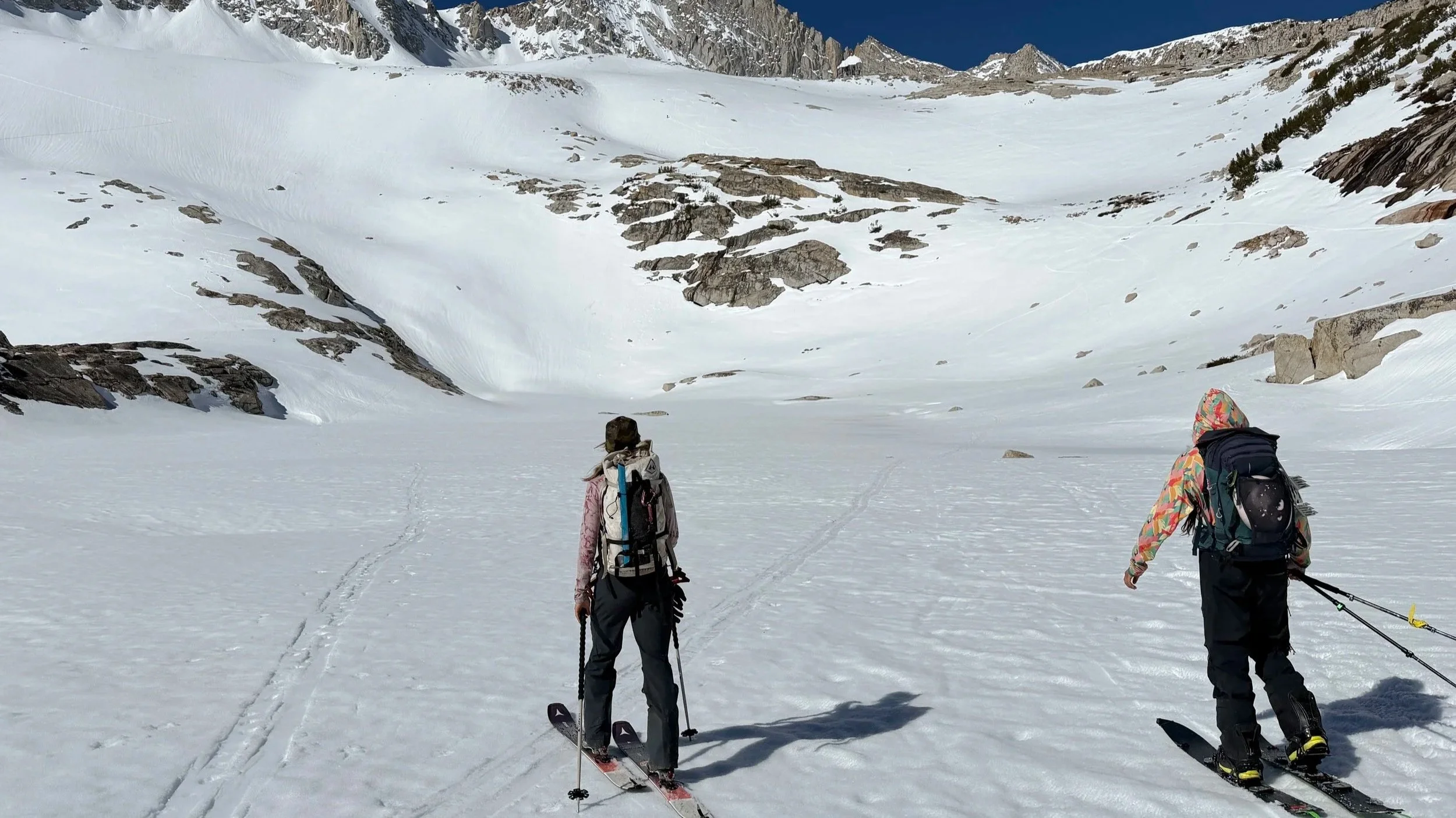 Two people wearing winter clothing and backpacks are skiing in a snow-covered mountain valley with rocky slopes and a blue sky in the background.