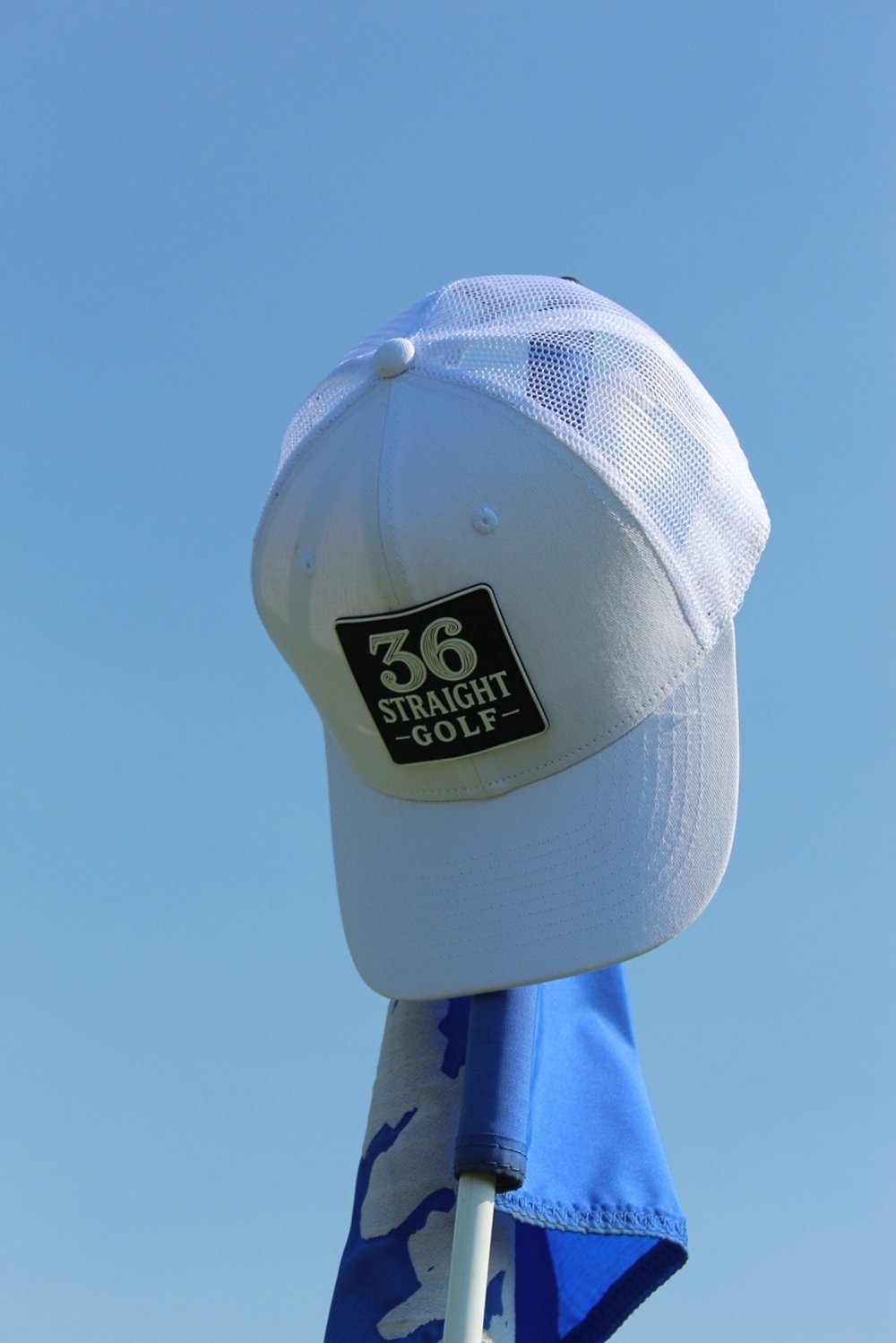 A golf hat with a black patch that reads "36 Straight Golf," mounted on a stand with a blue and white towel hanging from it, against a blue sky background.