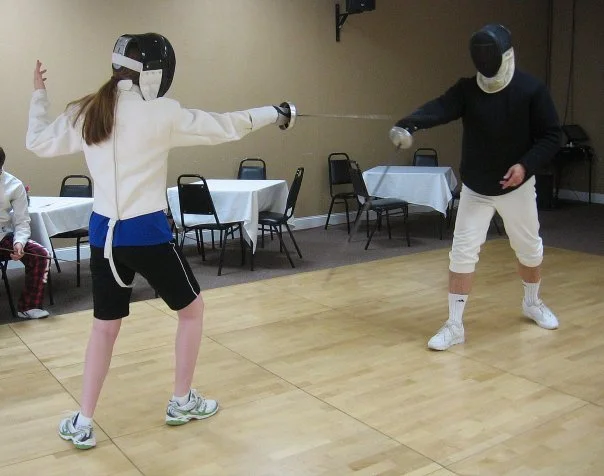 Two individuals in fencing gear practicing fencing indoors, with one person lunging at the other. There are empty chairs and tables in the background.