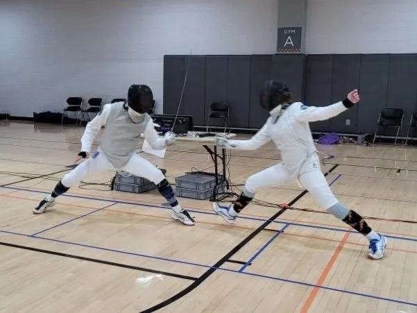Two people practicing fencing in a gym, wearing protective gear including masks and jackets, with fencing swords, while sparring.