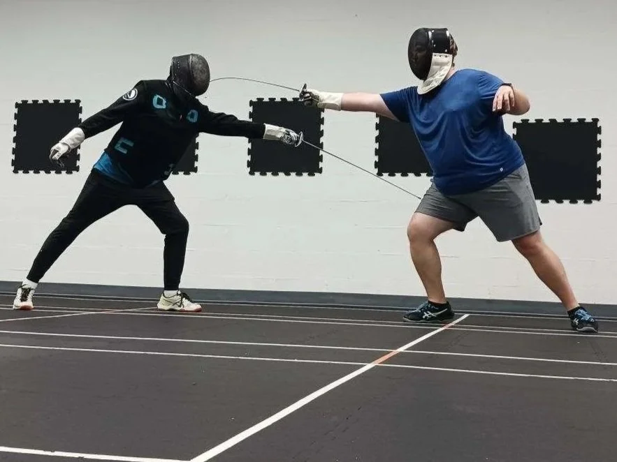 Two people engaged in a fencing match practicing on an indoor sports floor, both wearing protective masks and gear.