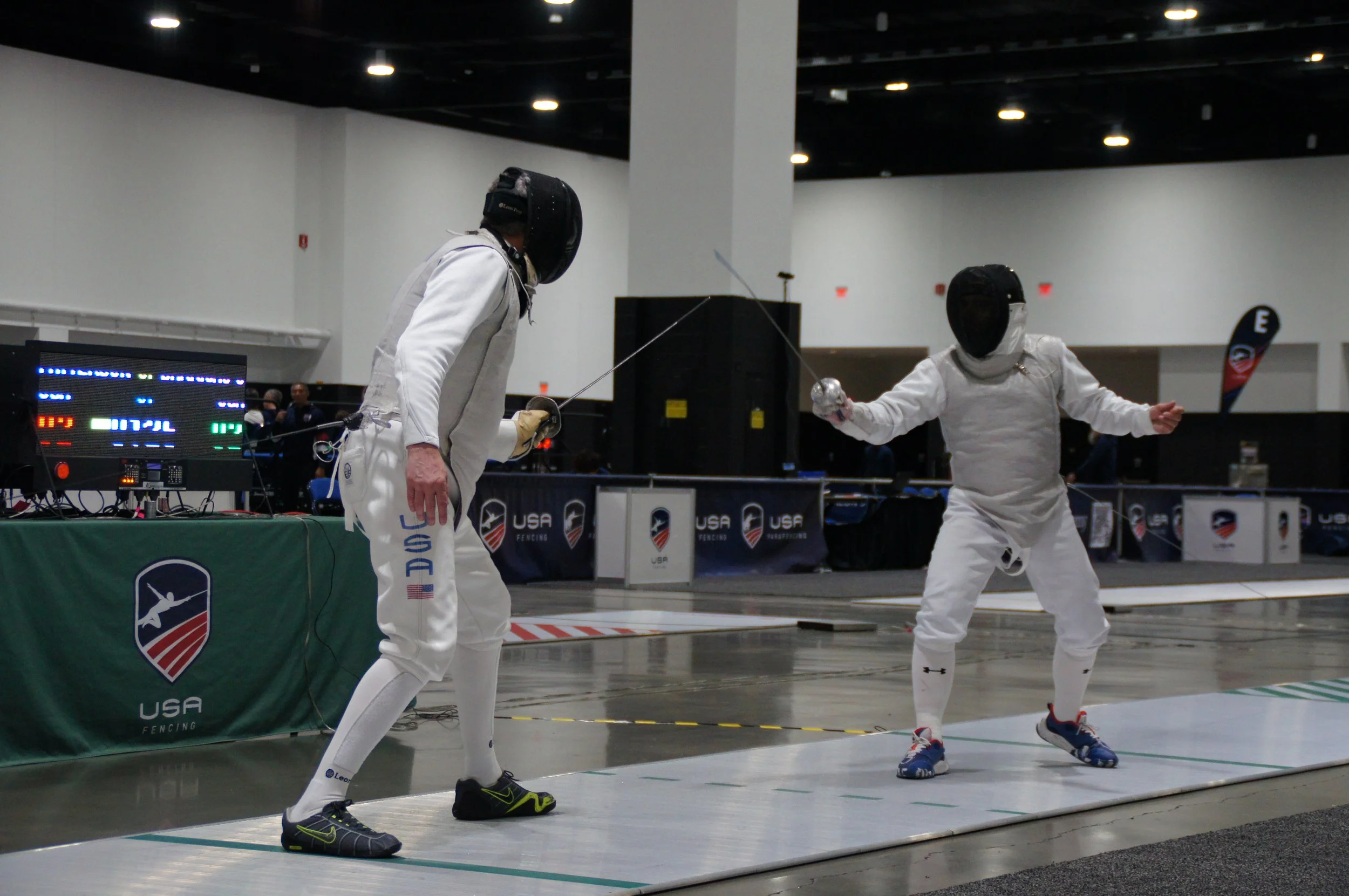 Two fencers in white protective gear and black masks engaging in a fencing bout at an indoor USA fencing event, with electronic scoring equipment nearby.