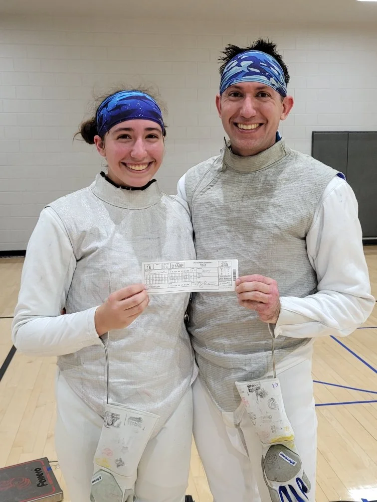 Two people wearing fencing uniforms and blue head coverings, holding a fencing match score sheet, smiling in a gymnasium.