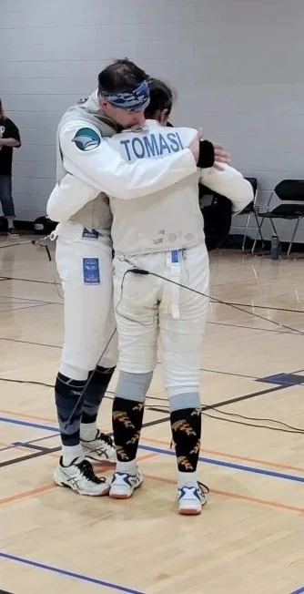 Two athletes in fencing gear embracing in a gymnasium.