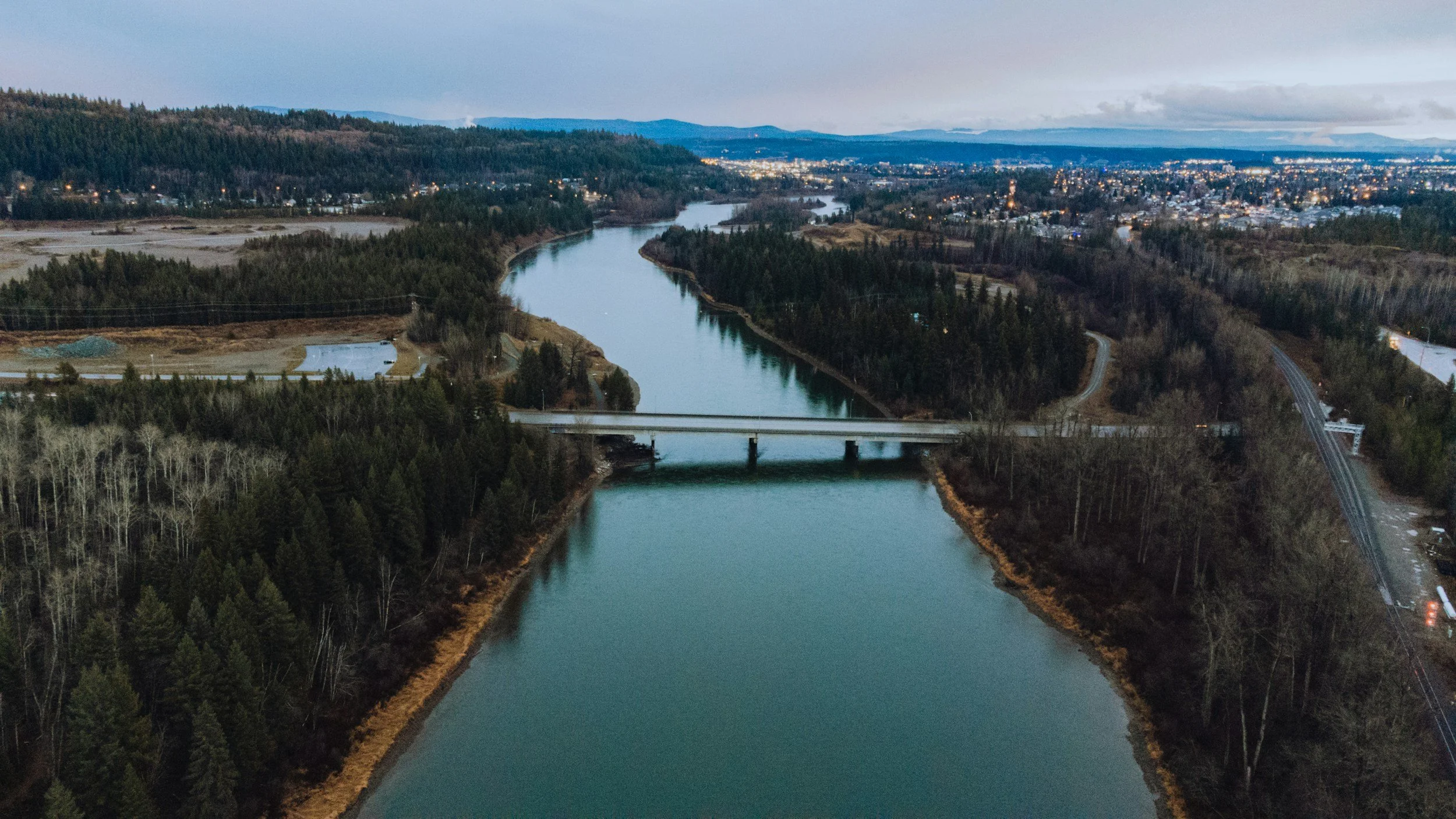 Aerial view of a river flowing through a forested landscape with a bridge crossing over it and a city in the distance under a cloudy sky.