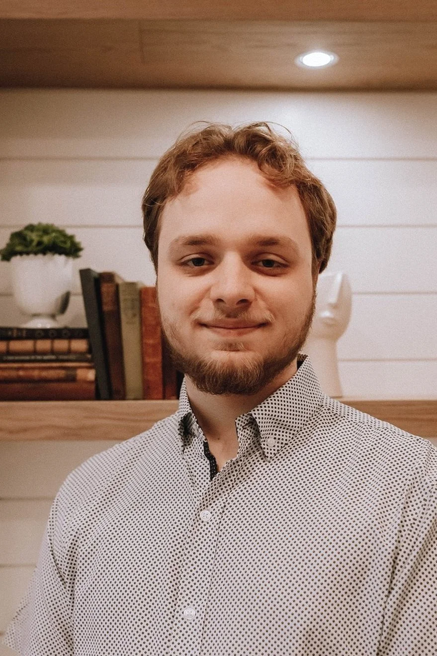 A young man with light brown hair, beard, and mustache, wearing a white shirt with small black dots, standing indoors in front of a white wall with wooden shelves, books, and a decorative white bust.