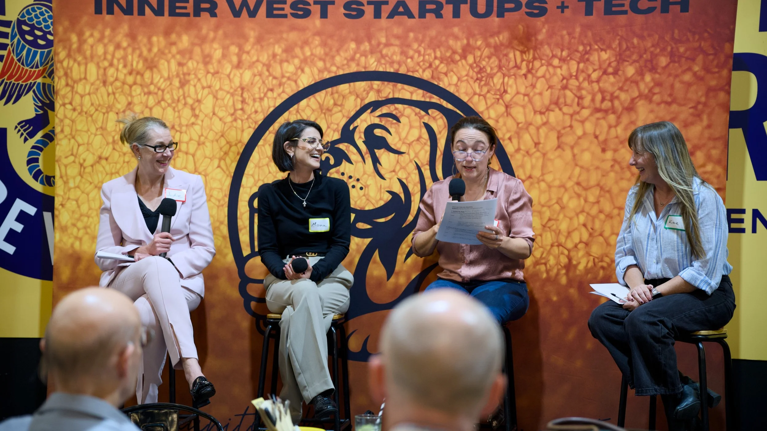 Four women sitting on stage at a panel discussion at an event called Inner West Startups + Tech, with a large orange backdrop featuring black text and a stylized eagle logo. The women are smiling, holding microphones or notes, and are engaged in conversation.