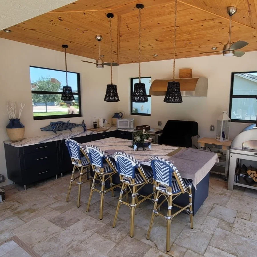 A outdoor kitchen with a wooden ceiling, blue cabinets, and a large island with six chairs. There are pendant lights above the island, a large grill and pizza oven
