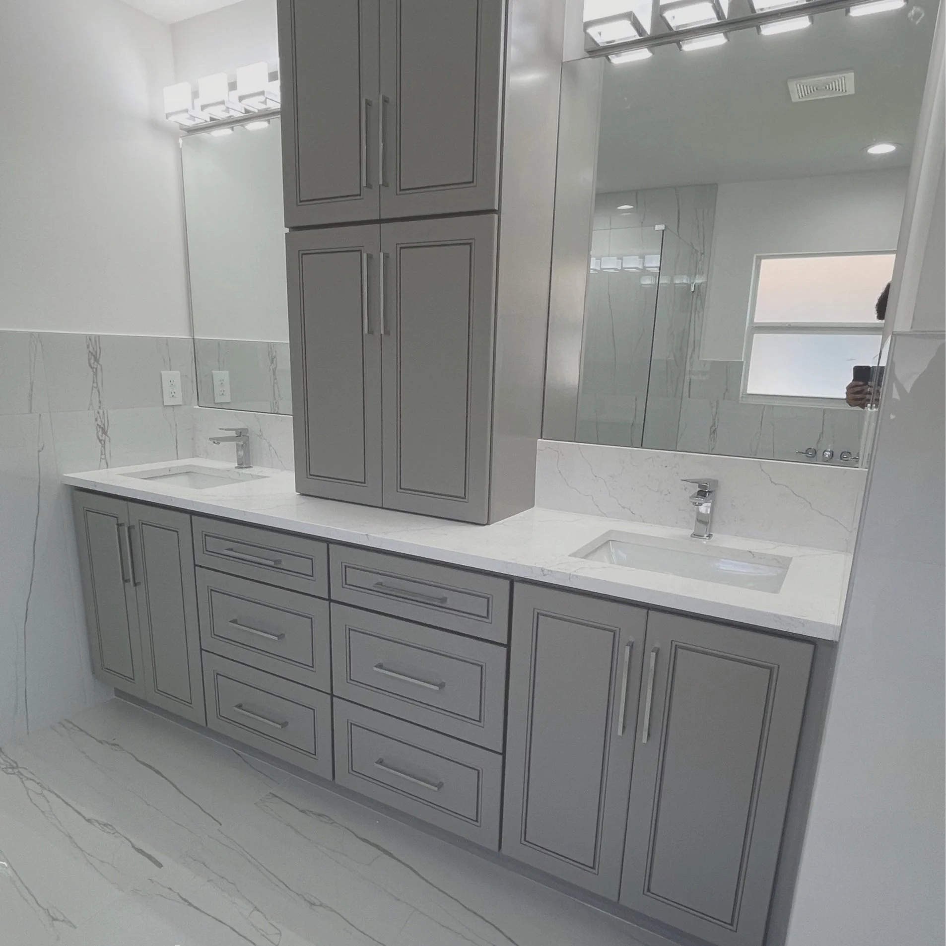 Modern bathroom vanity with gray cabinets and multiple drawers, a white countertop with two rectangular sinks, large mirror, and wall-mounted light fixtures, with a window in the background.