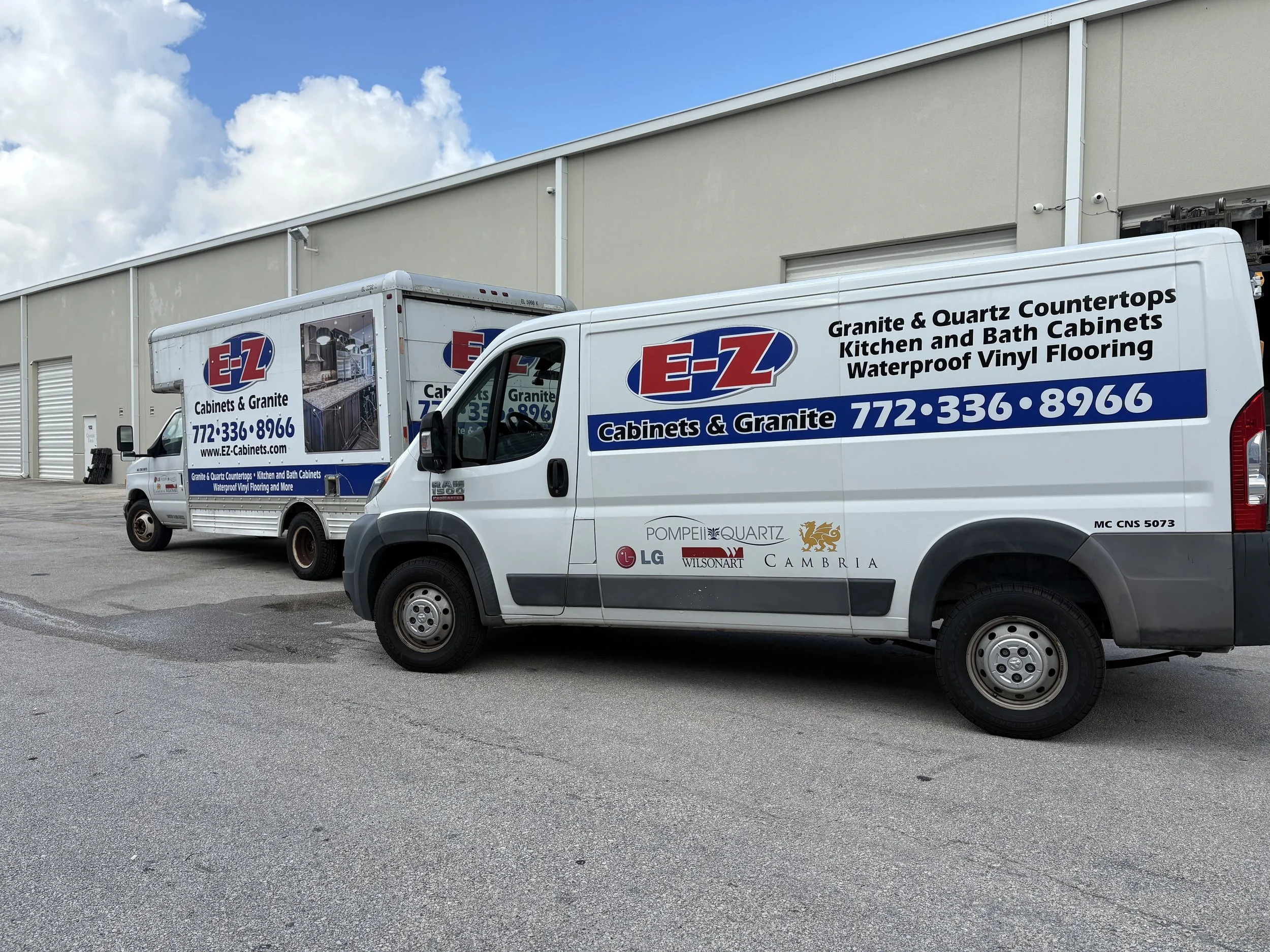 Two white delivery vans with 'E-Z Cabinets & Granite' branding parked outside a beige industrial building under a partly cloudy sky.
