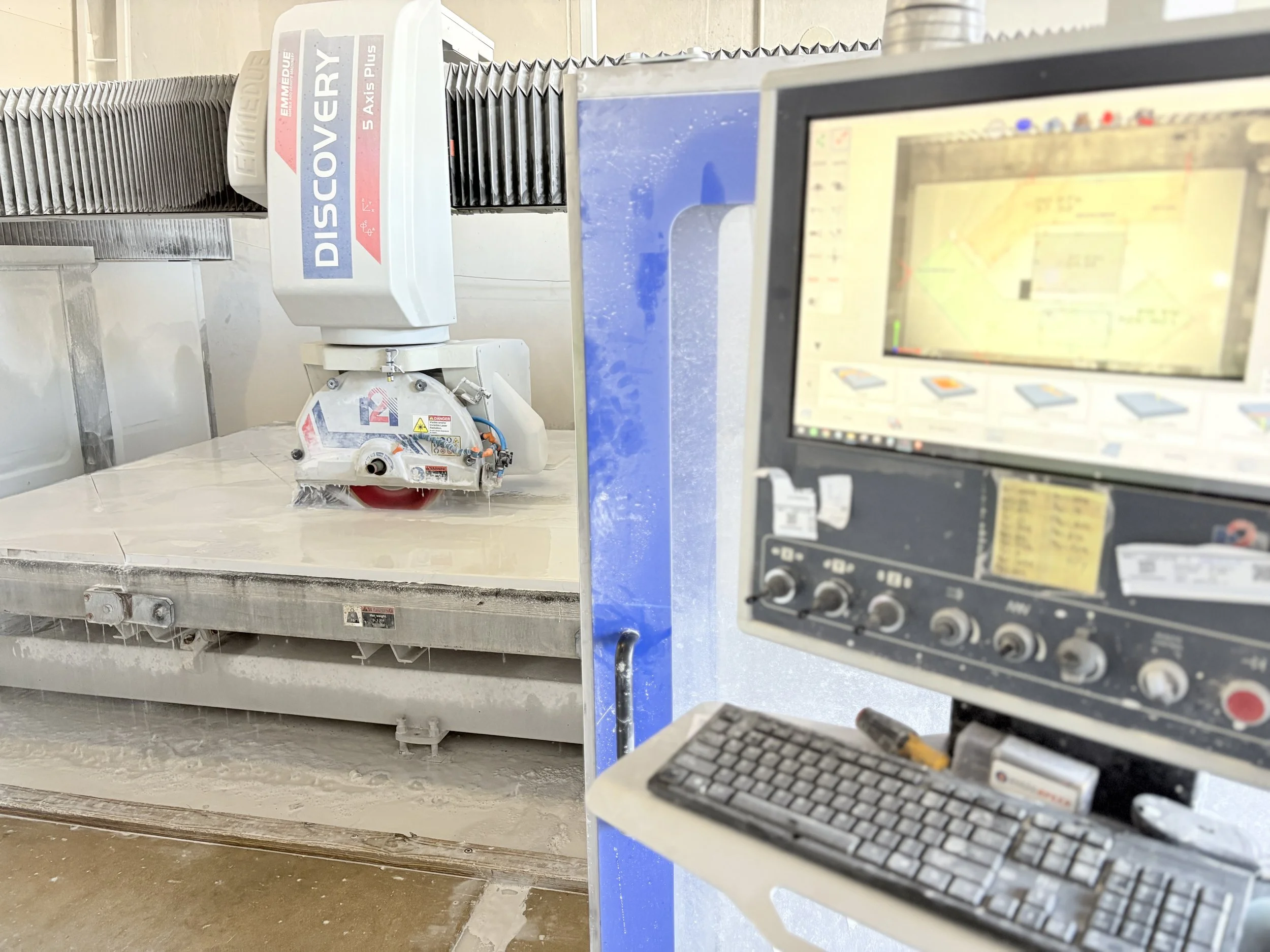 A large CNC machine cutting a stone slab with a mounted dust extraction system above it, and a computer monitor in the foreground displaying design specifications.
