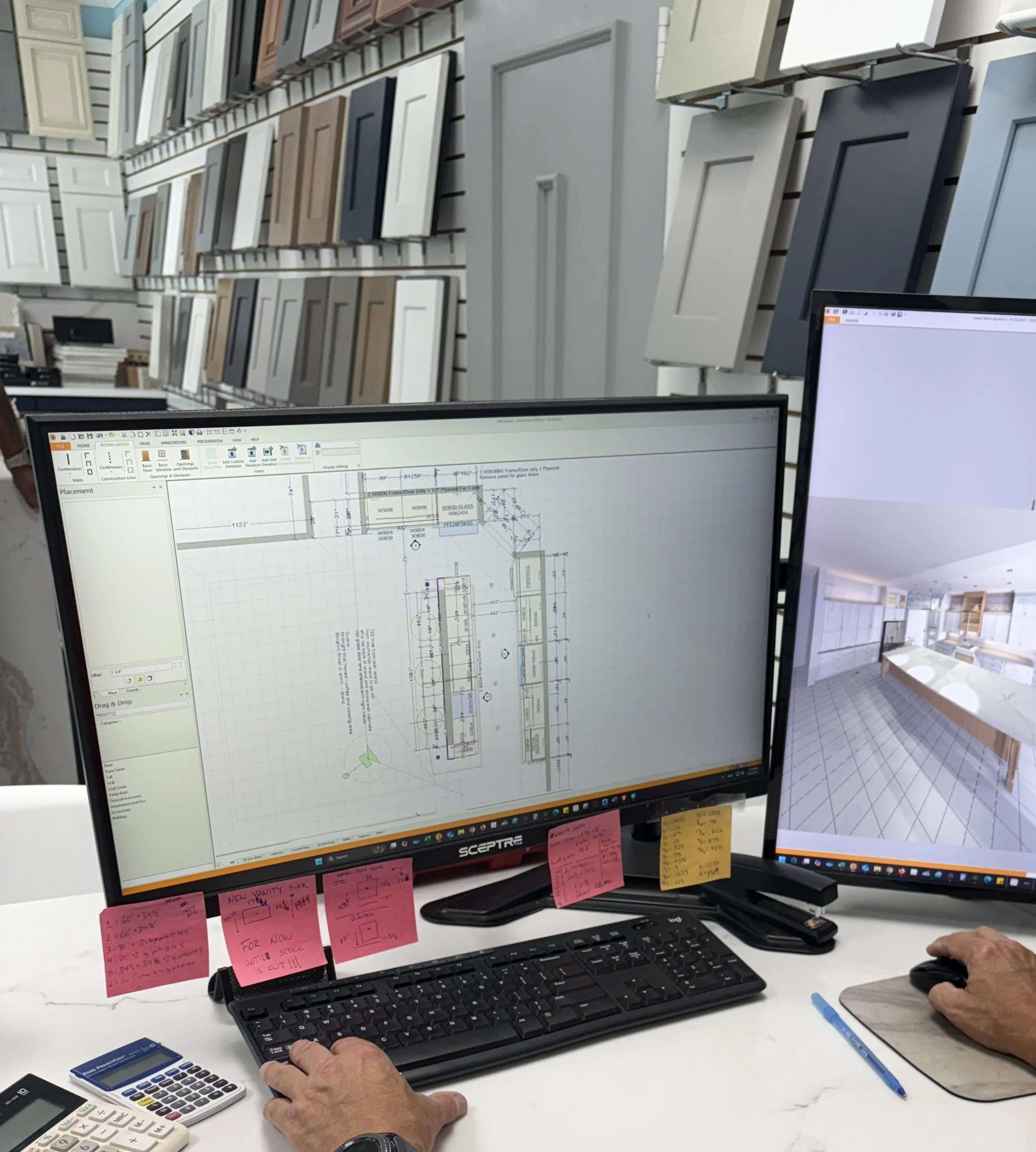 A person working on computer monitors displaying architectural floor plans and 3D renderings in an interior design or architecture office. Behind the monitors, sample cabinet doors are mounted on a slatwall, indicating home improvement or interior design context.
