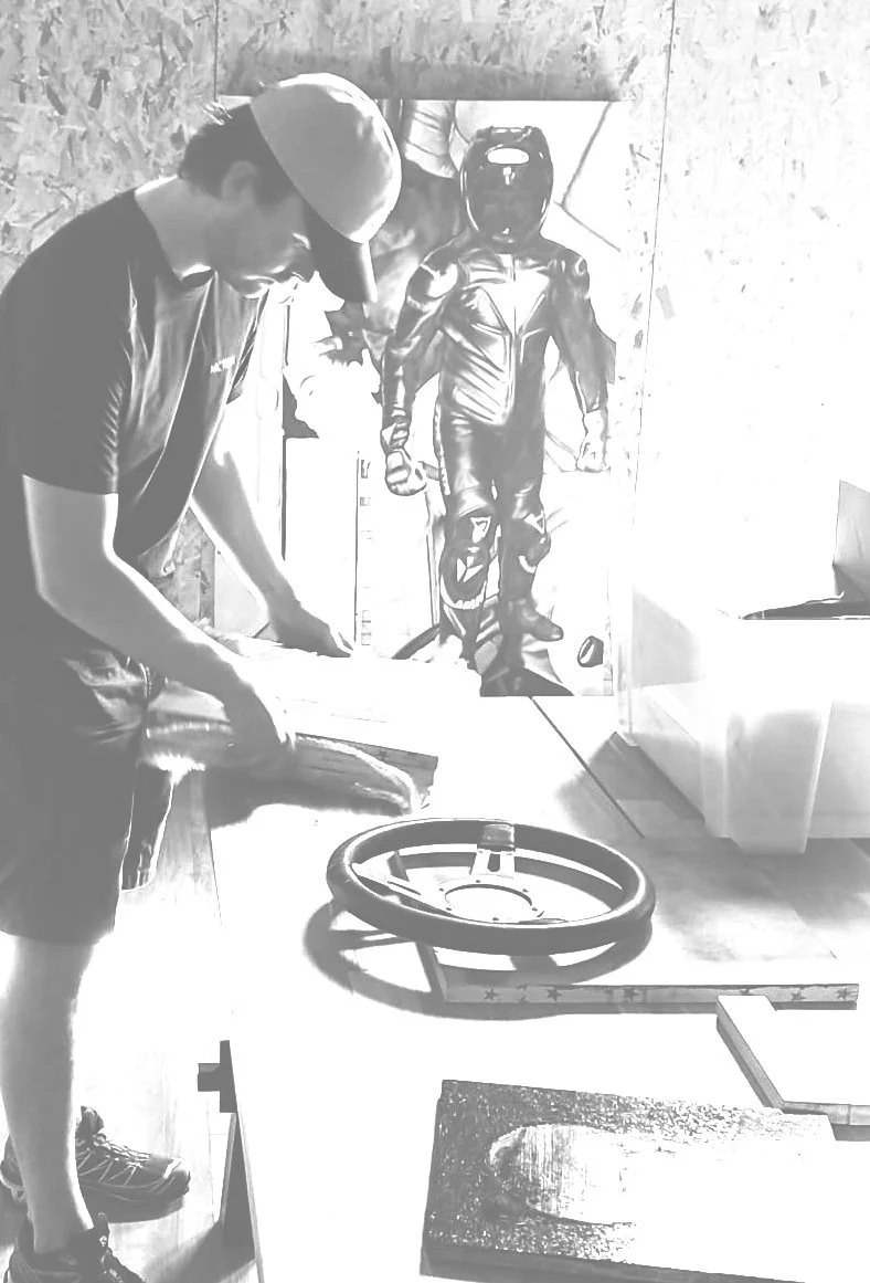 A young man with a cap looks down at a table with a wheel and tools, in front of a poster of a motorcyclist in racing gear.
