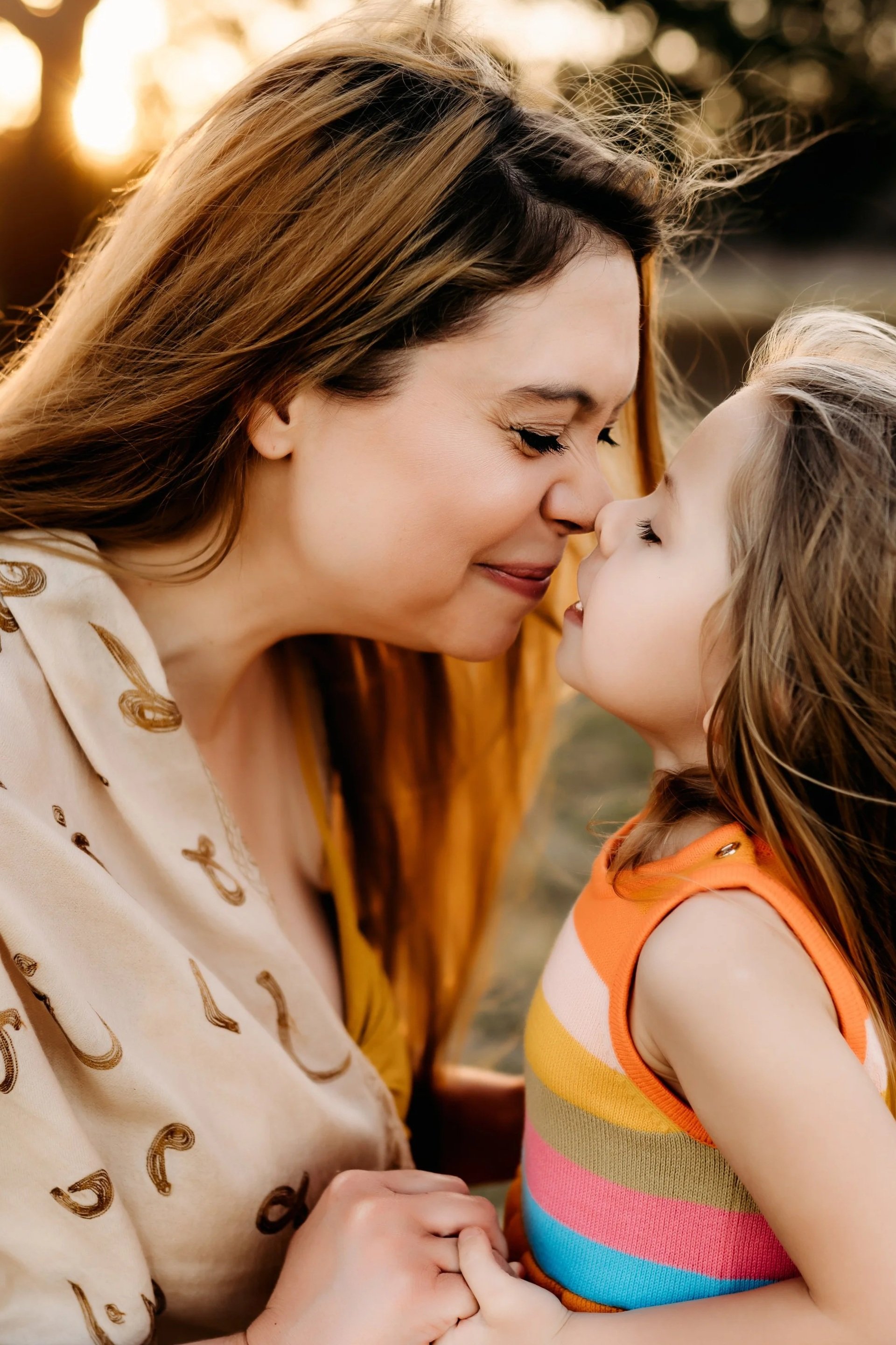 A woman and a young girl are touching noses and smiling at each other outdoors during sunset.