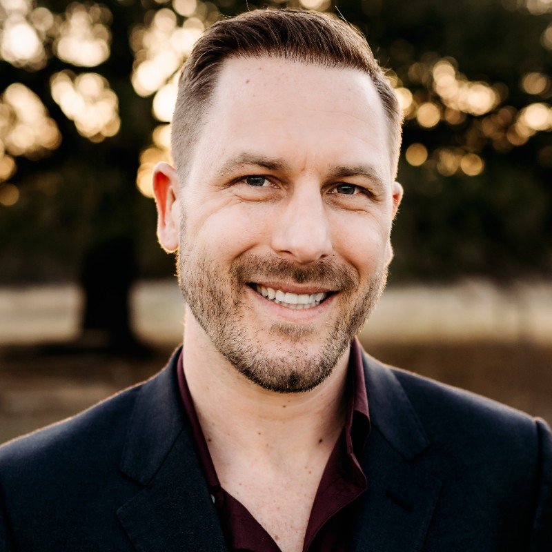 A smiling man with short brown hair and a beard, wearing a dark blazer and maroon shirt, standing outdoors during sunset with blurred trees in the background.
