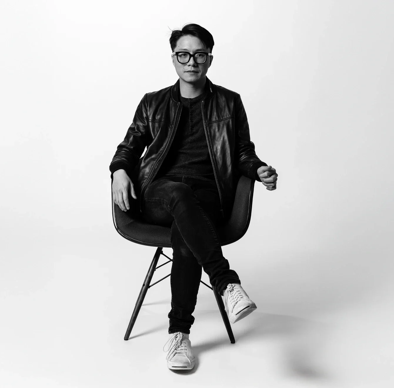 Black and white photo of a man sitting on a chair against a plain background, wearing glasses, a leather jacket, dark clothing, and sneakers.