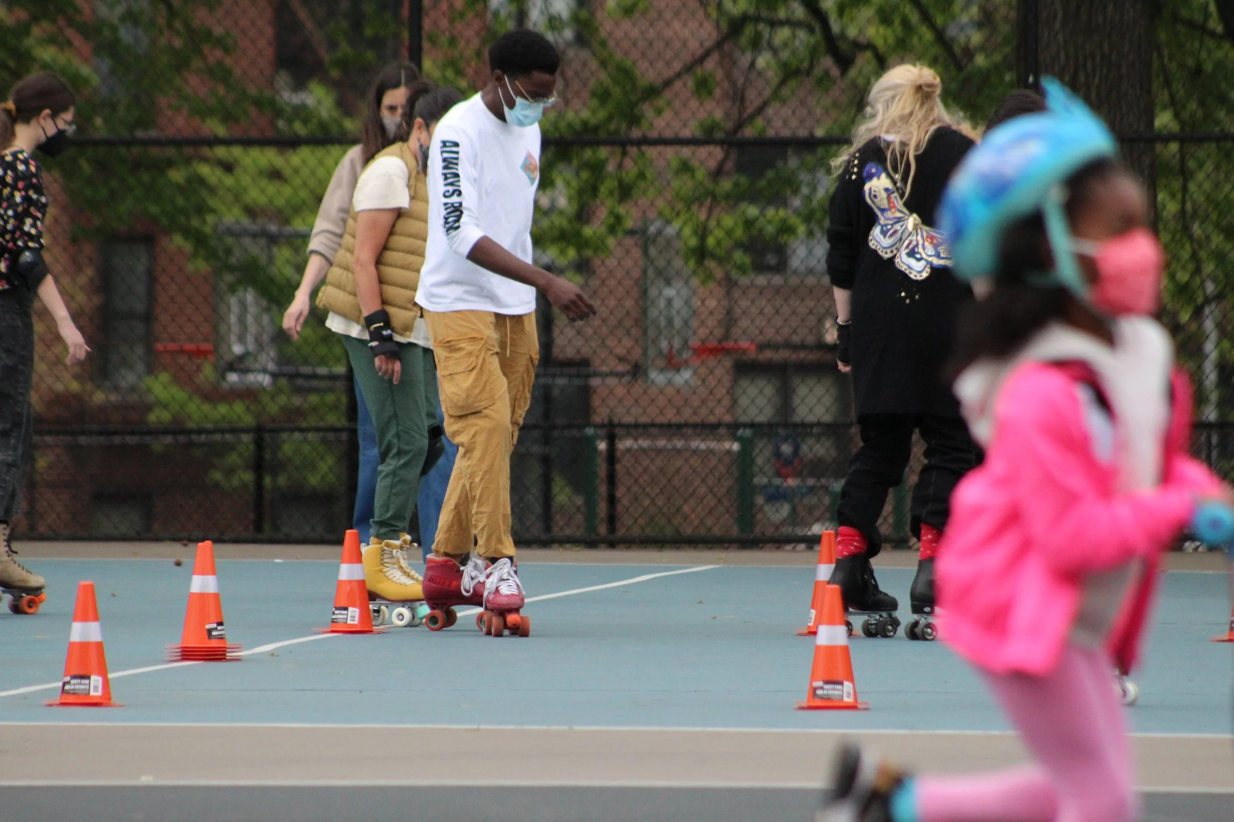 People roller skating on an outdoor court, practicing skating skills with orange traffic cones set up as obstacles. The skaters are wearing masks and casual clothing, with a chain-link fence and trees in the background.