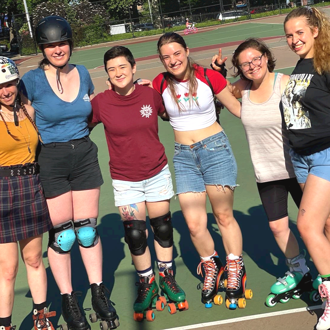 Six women standing together with their arms around each other on a roller skate rink outdoors, smiling at the camera on a sunny day.