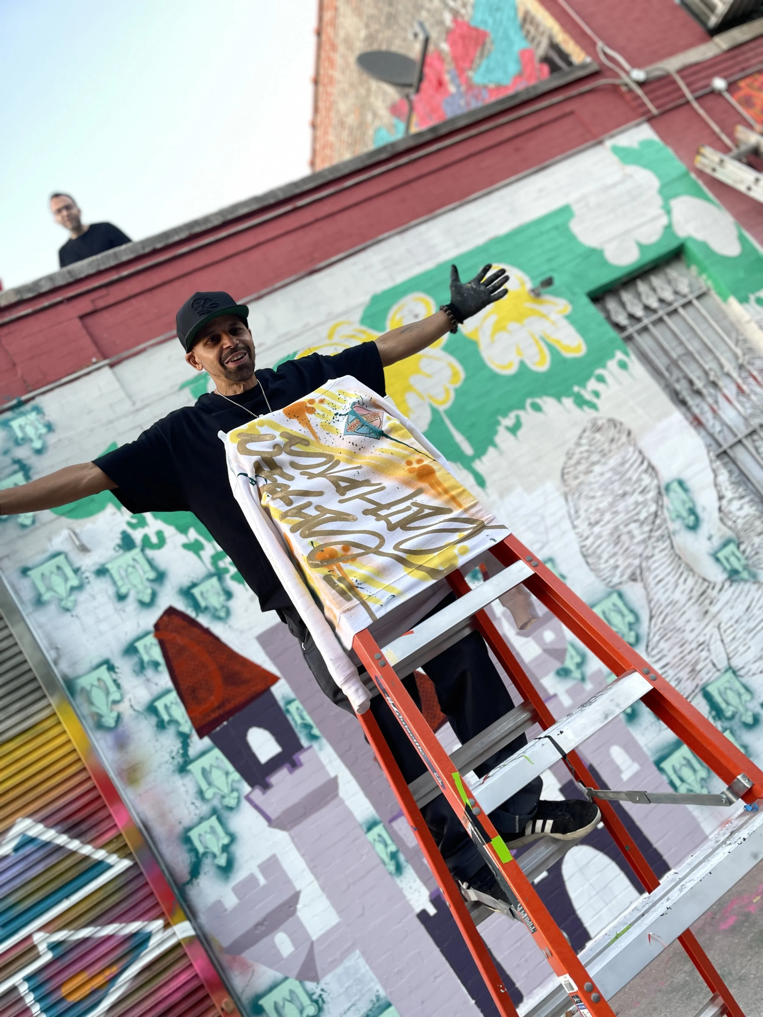 A man wearing a black cap, black shirt, and black gloves stands on a red ladder in front of a colorful mural with abstract designs and fish on a brick wall. Another person is visible on the roof in the background.