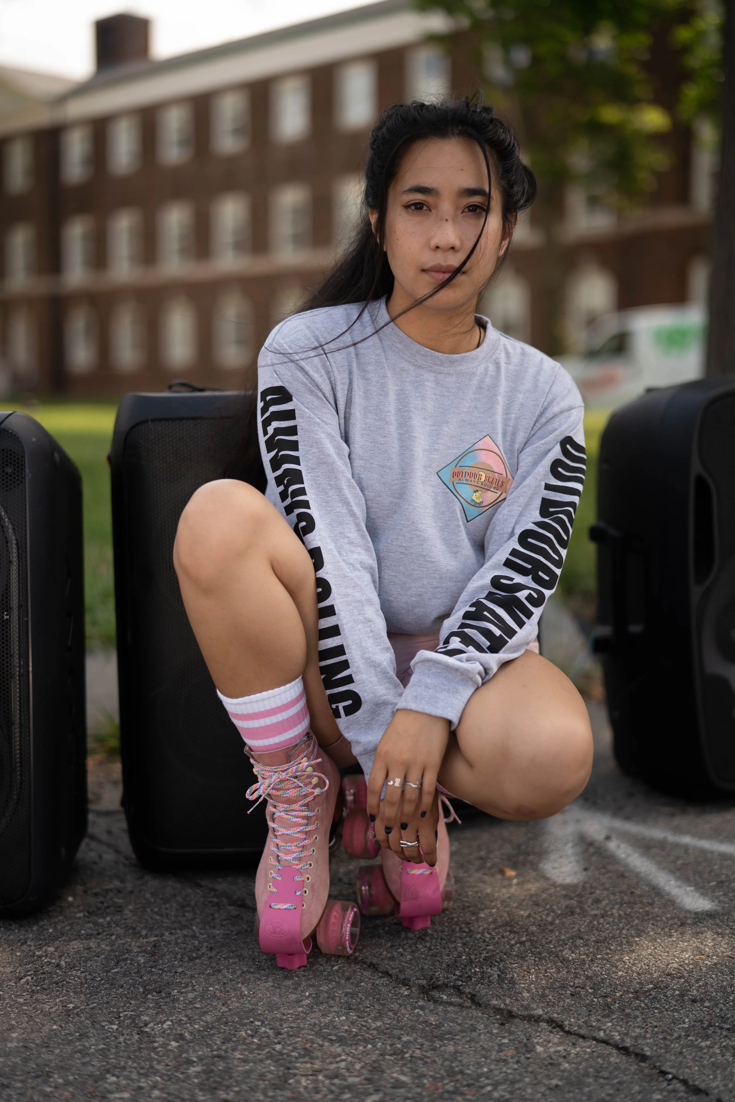 Young woman with long dark hair and freckles sitting on the ground with roller skates on, outdoors on a paved area with a background of a large brick building, some trees, and a parked van.
