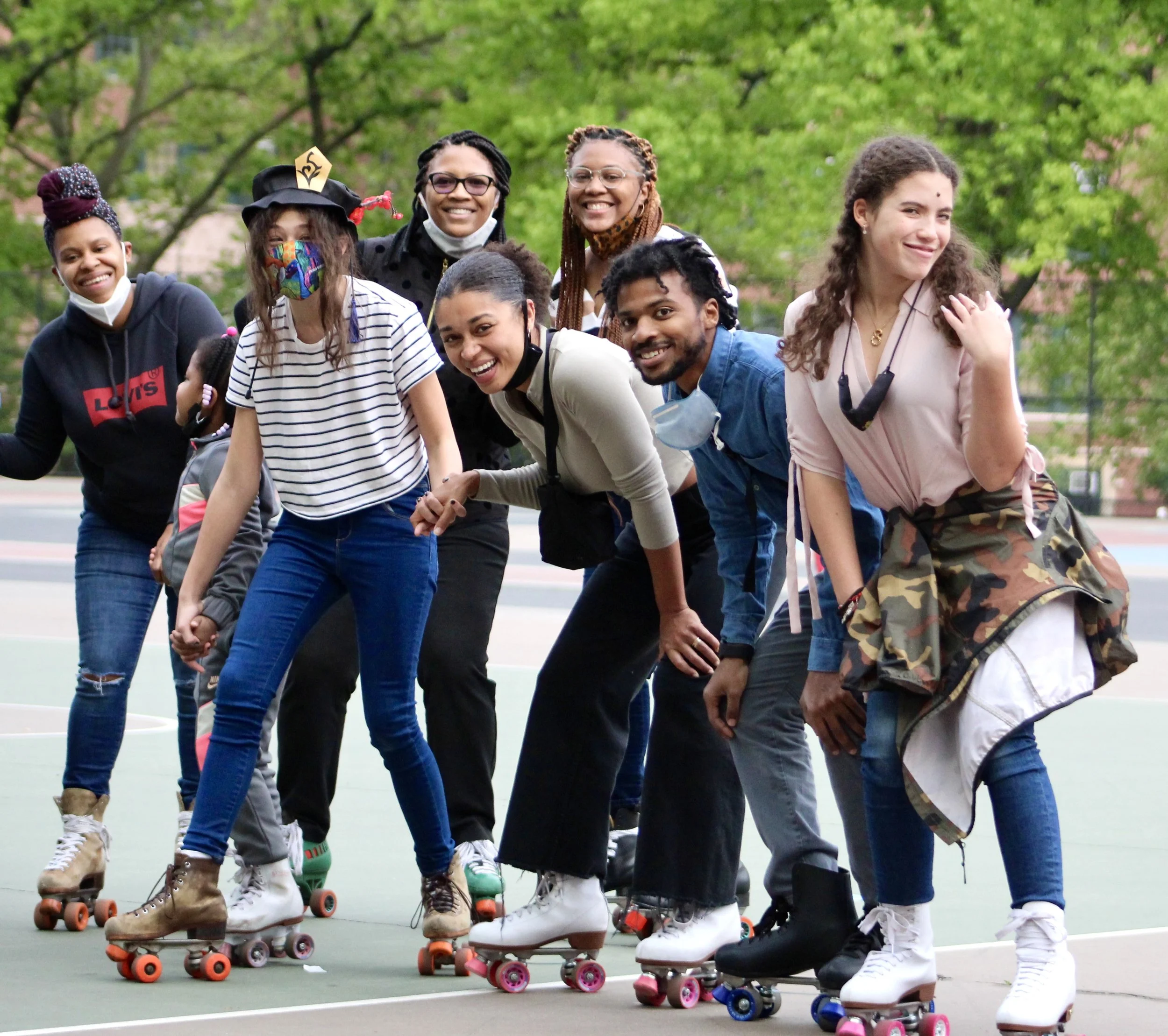 A group of diverse people on roller skates in a line, smiling and holding hands outdoors with green trees in the background.