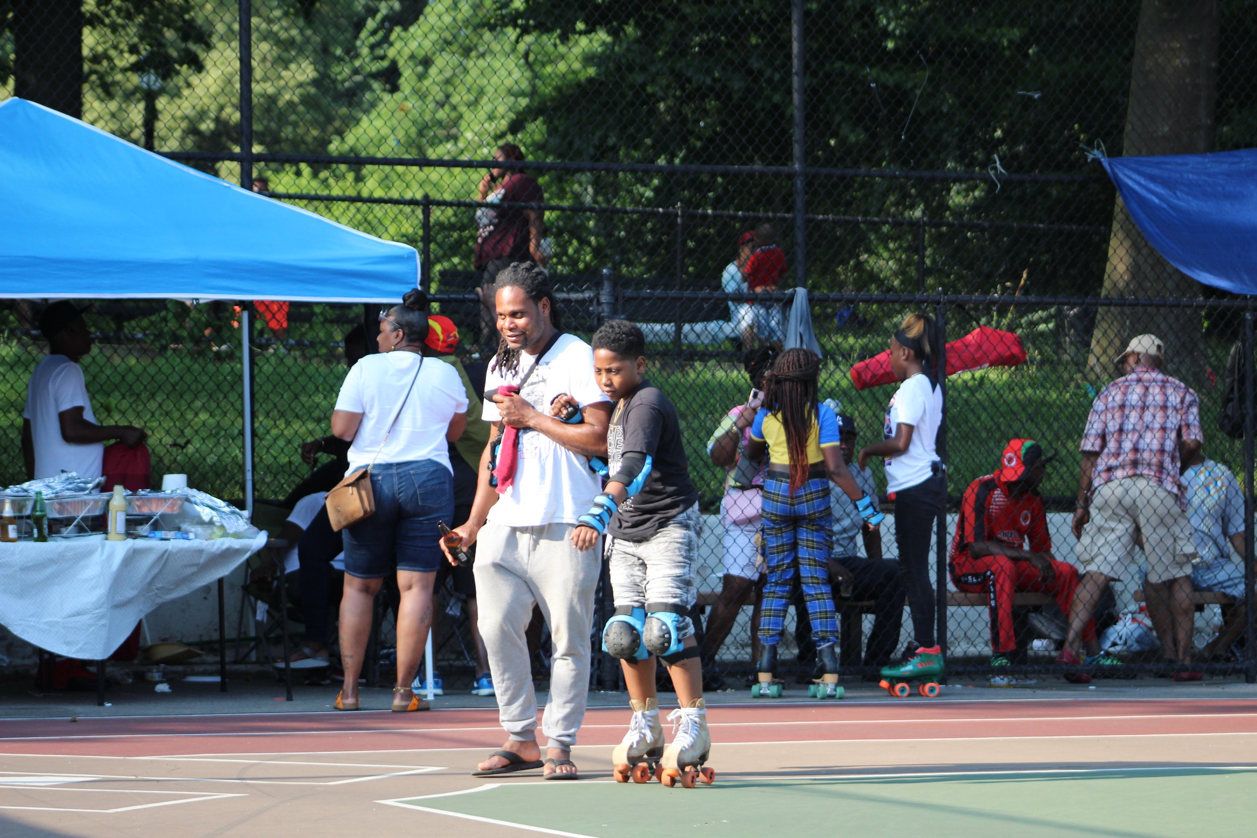 People at an outdoor skate event near a chain-link fence, with trees in the background. Some are roller skating and others are watching or talking.