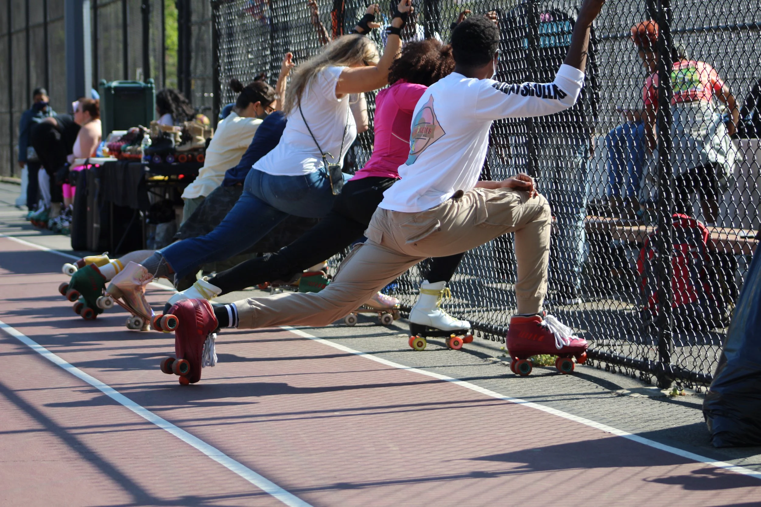 People roller skating outside on a sports court beside a chain-link fence, with some sitting and others standing or skating along the fence, during daylight.