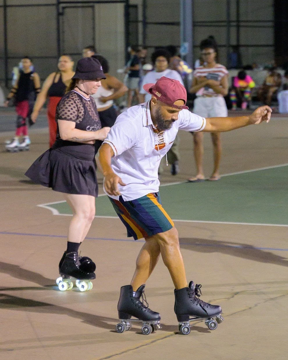 People roller skating on an outdoor court at night, including a man with a beard in a white shirt and colorful shorts, and a woman with a black hat and a black dress.