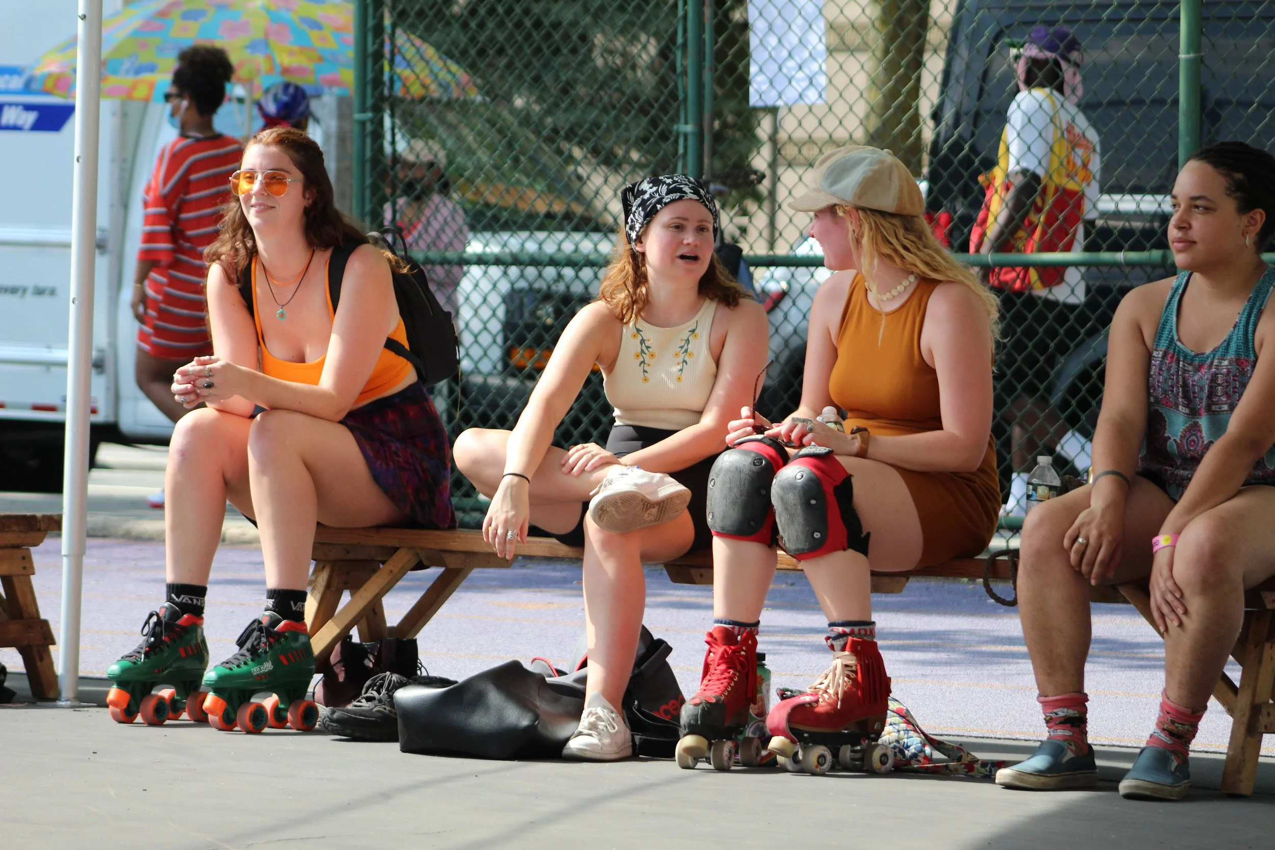 A group of young women sitting on a wooden bench at an outdoor skating rink, with roller skates and protective gear on them. They are talking and relaxing in a casual summer setting.