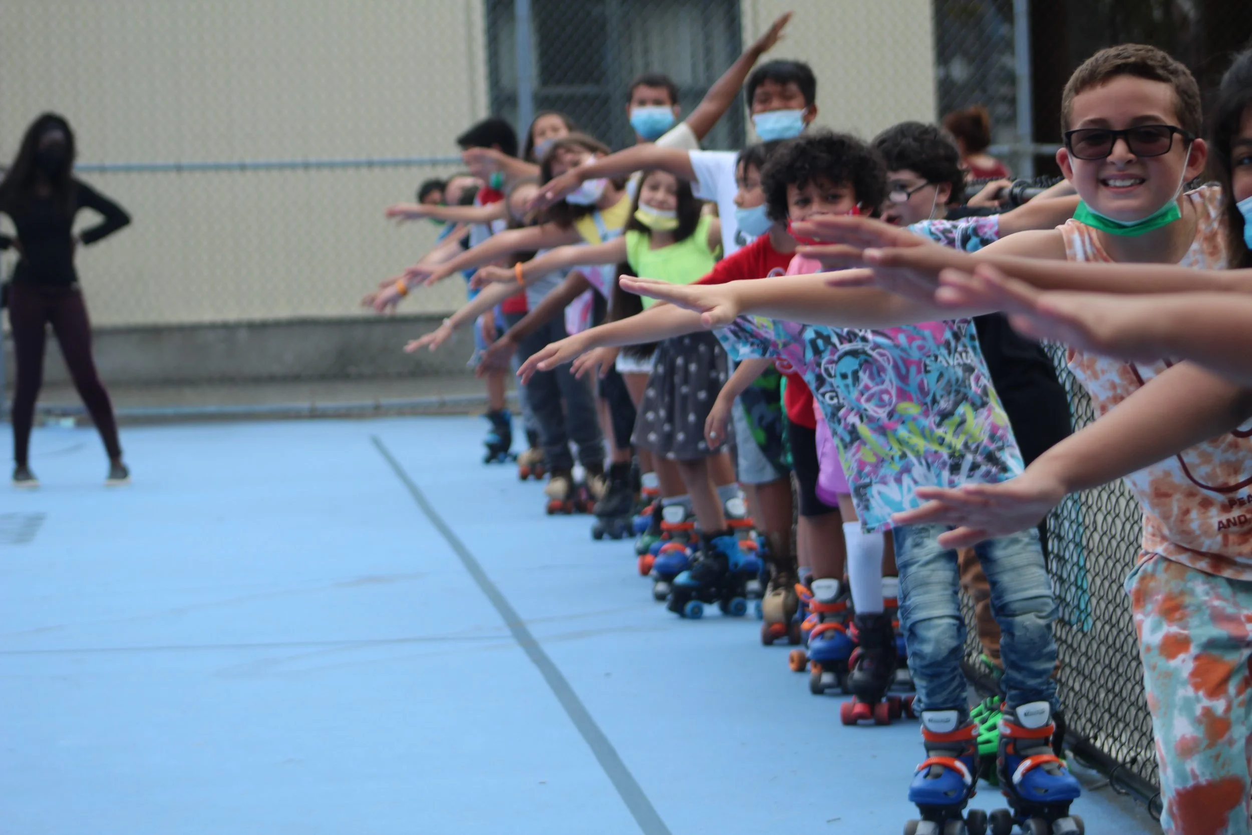 Children wearing masks and roller skates line up on a sports court, extending their arms forward for a skating activity. An instructor stands to the side.