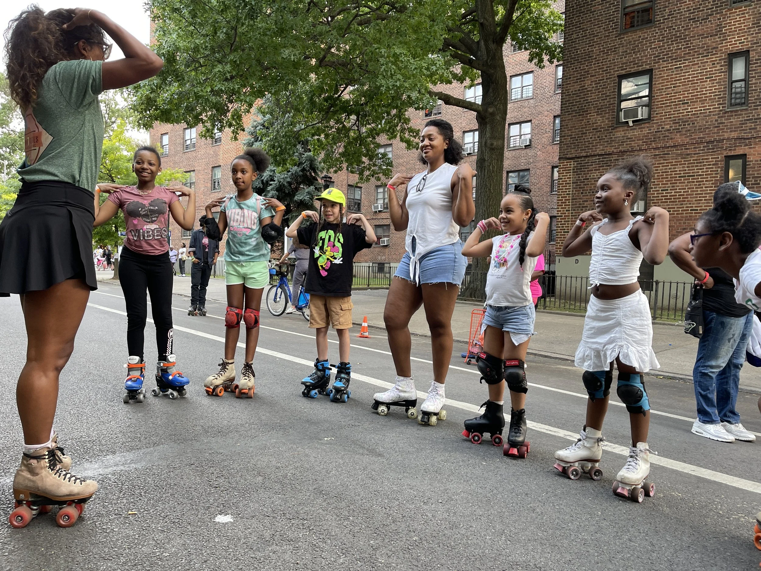 A group of young girls with roller skates lined up on a city street, wearing protective gear like helmets and knee pads, attending a roller skate lesson, in front of brick apartment buildings and trees.