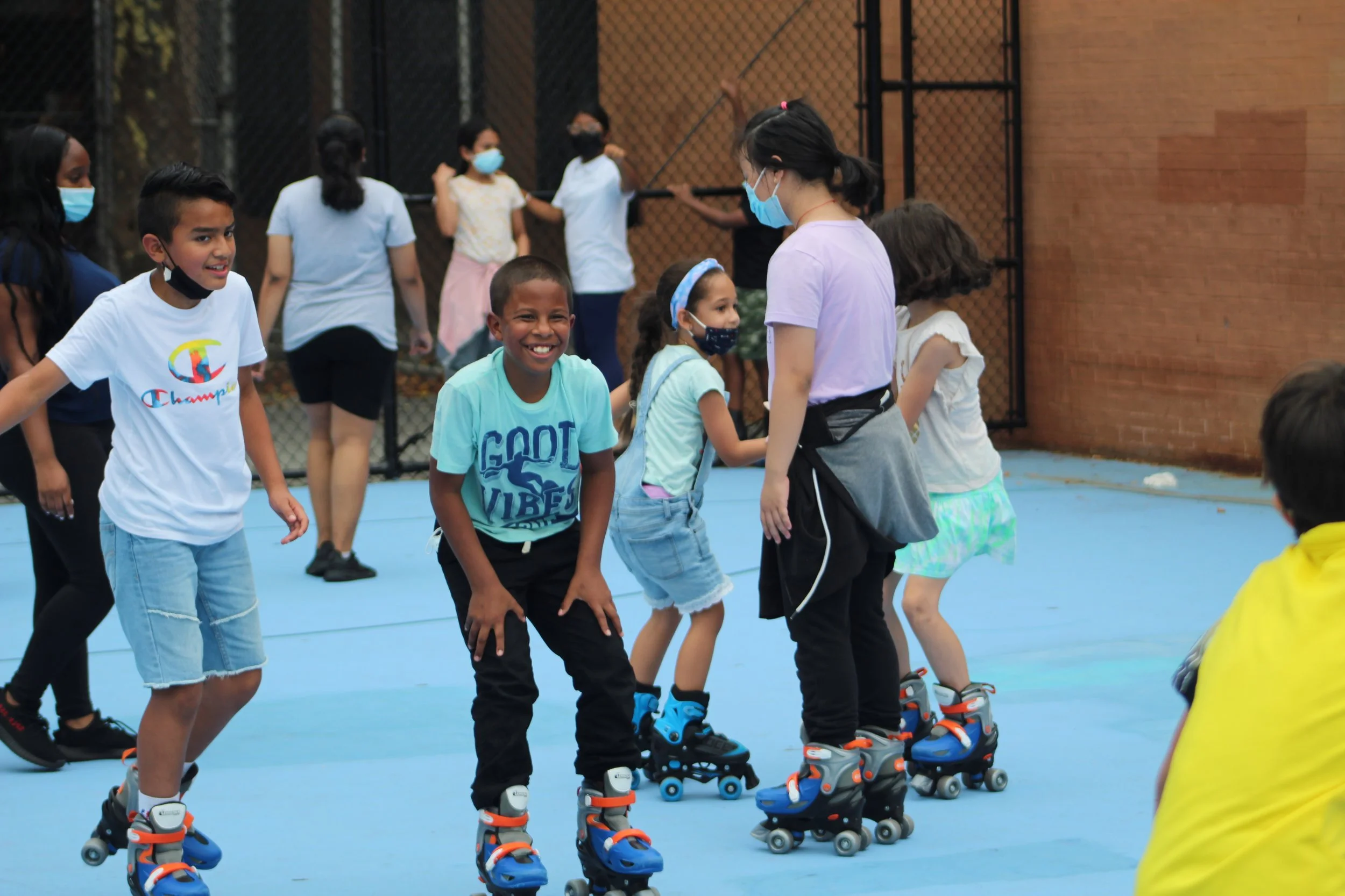 Children wearing face masks roller skating outdoors on a blue court.