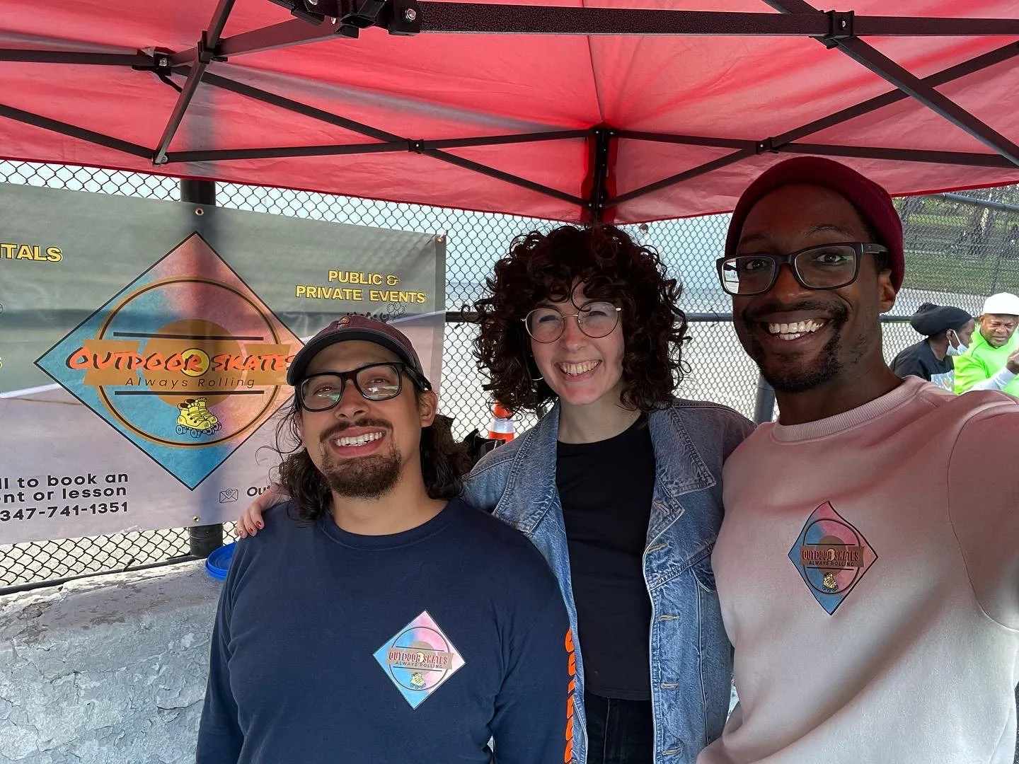 Three smiling people taking a selfie under a red canopy at an outdoor event, with a banner that says "Outdoor Skates, Always Rolling" and some individuals in the background.