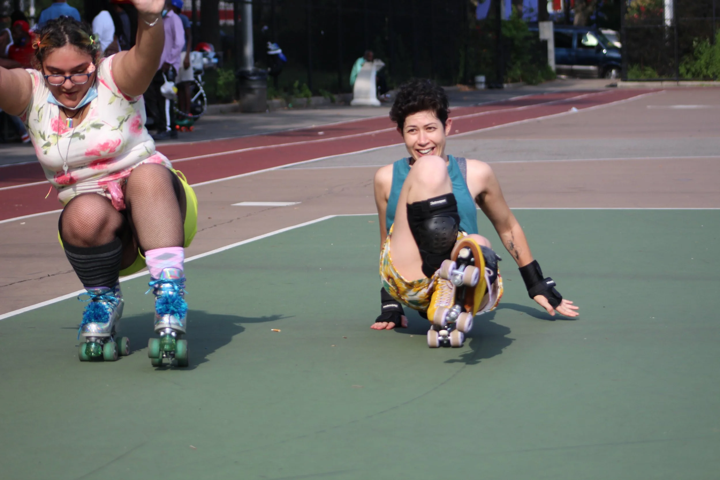 Two girls roller skating on an outdoor court, one dressed in a floral dress, and the other in a sleeveless top and shorts, who has fallen and is sitting on the ground smiling.