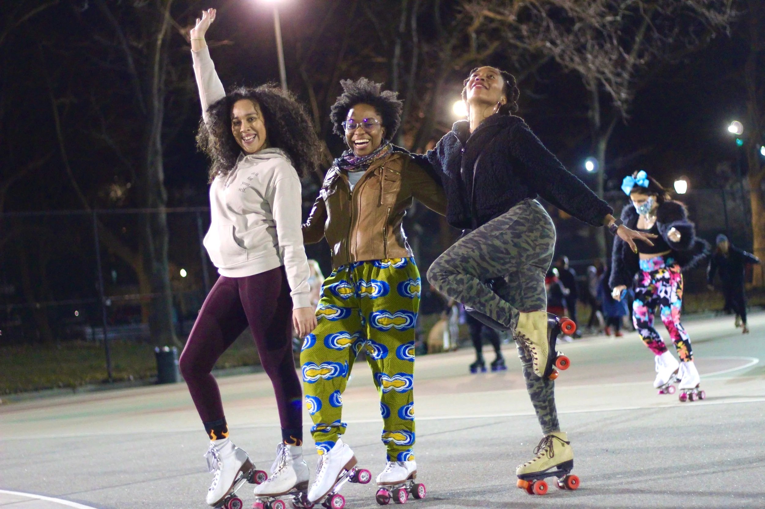 Three women roller skating at night on an outdoor basketball court, smiling and enjoying themselves. Two women are in front, and one is in the background wearing a face mask and colorful clothing.
