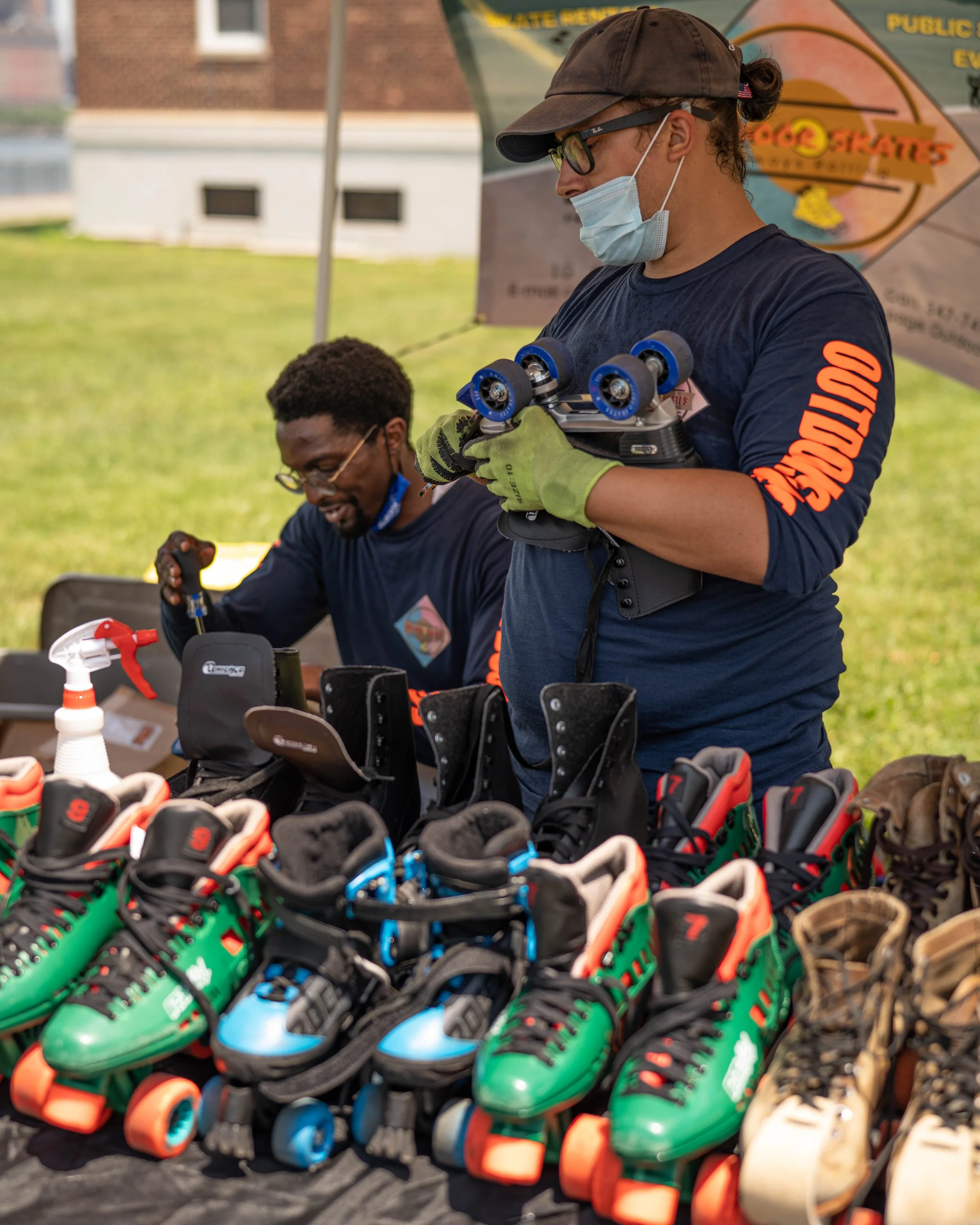 A person wearing a face mask, glasses, and a brown cap, holding roller skates, standing in front of a row of colorful roller skate shoes displayed on a table. There is another person in the background, also wearing glasses, engaged in an activity at the table, with a banner and building visible behind them.