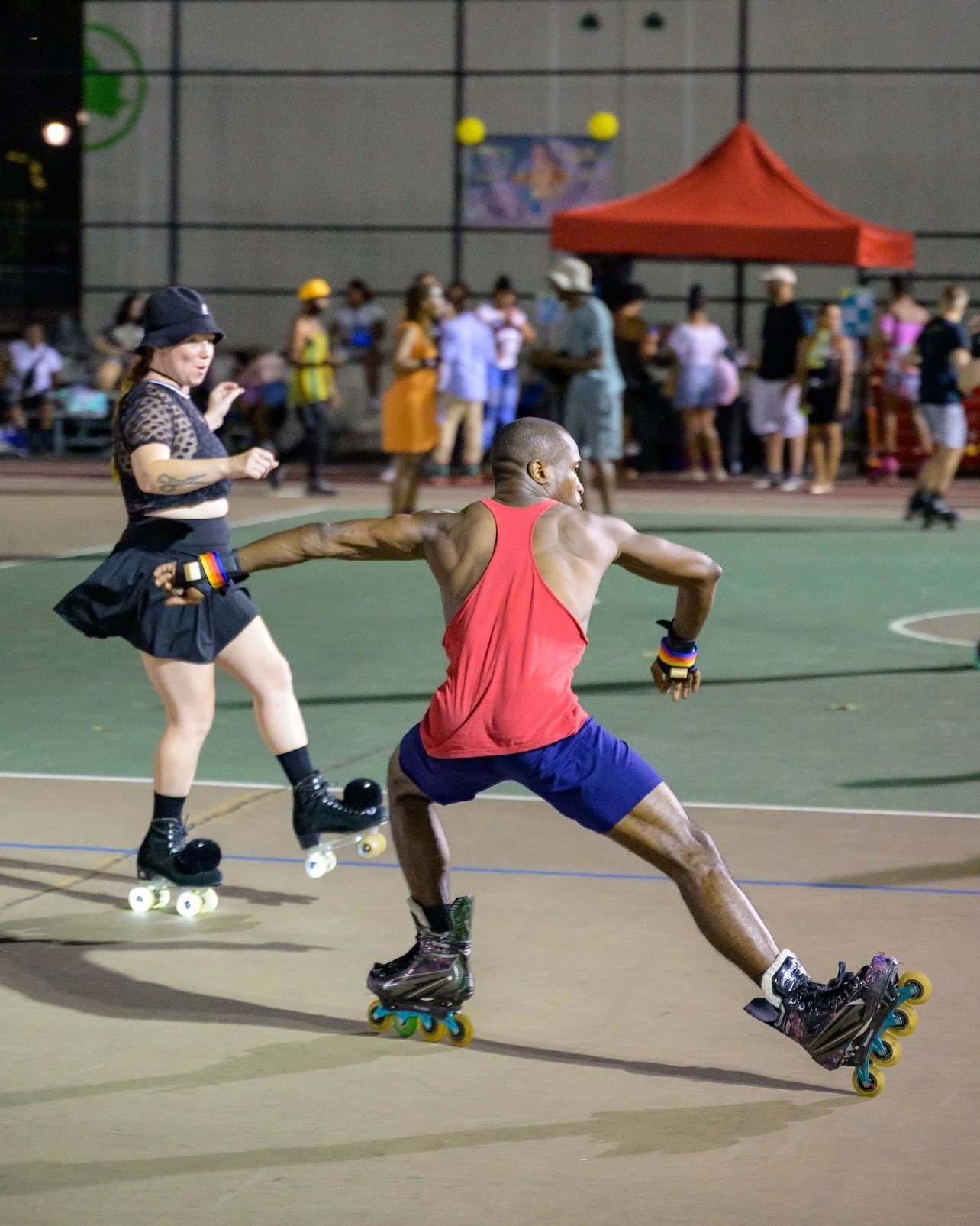 Two skaters in a roller derby match on an indoor court, with spectators in the background, one wearing a black hat and the other in a red tank top and blue shorts.