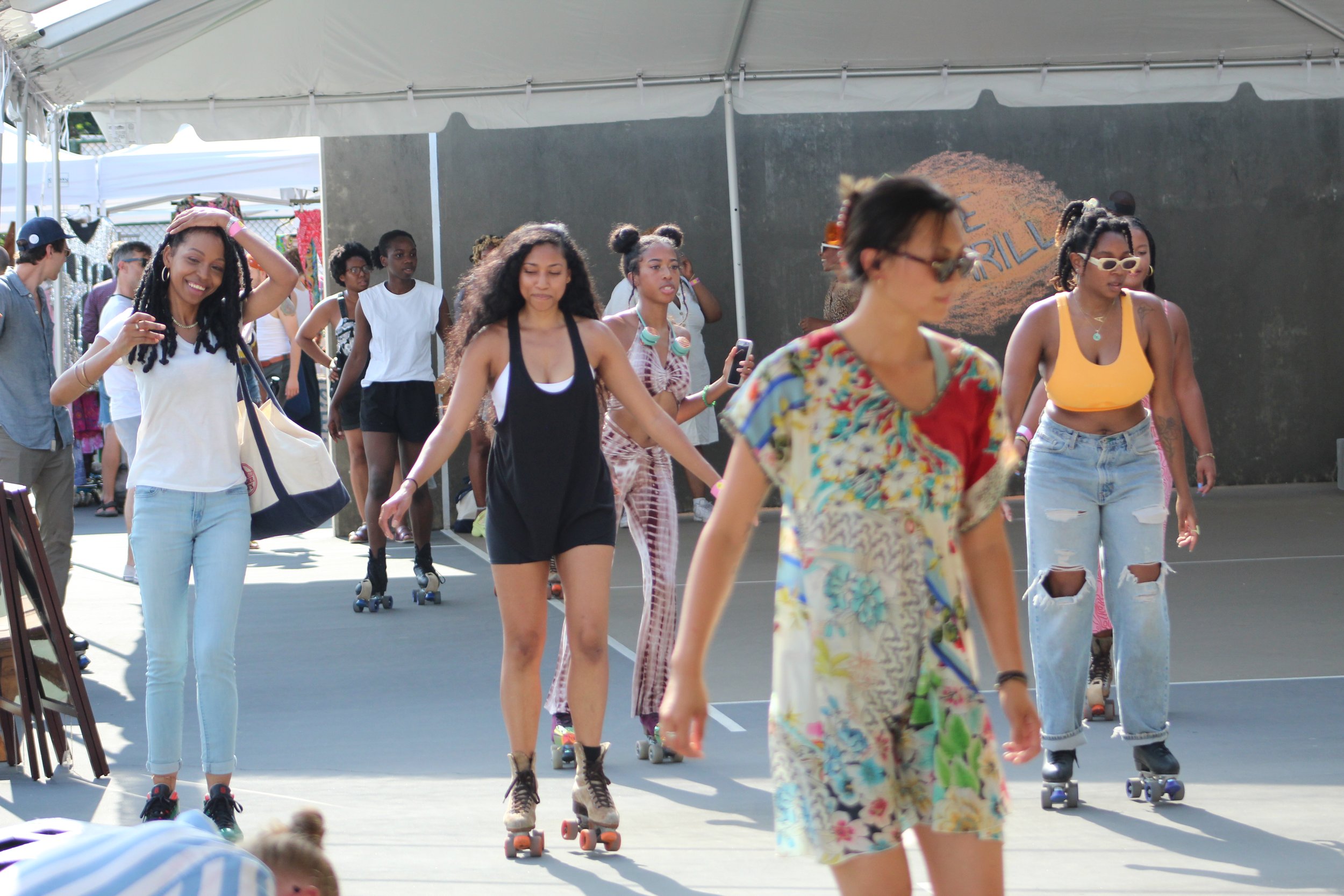 A group of young women and girls roller skating under a large tent at an outdoor event. Some are wearing casual clothing, and a few are holding phones.