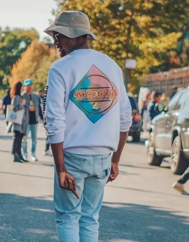 A person wearing a white sweatshirt with a colorful logo on the back, light blue jeans, a bucket hat, and sunglasses, standing on a street with cars and other people, with trees and a clear sky in the background.