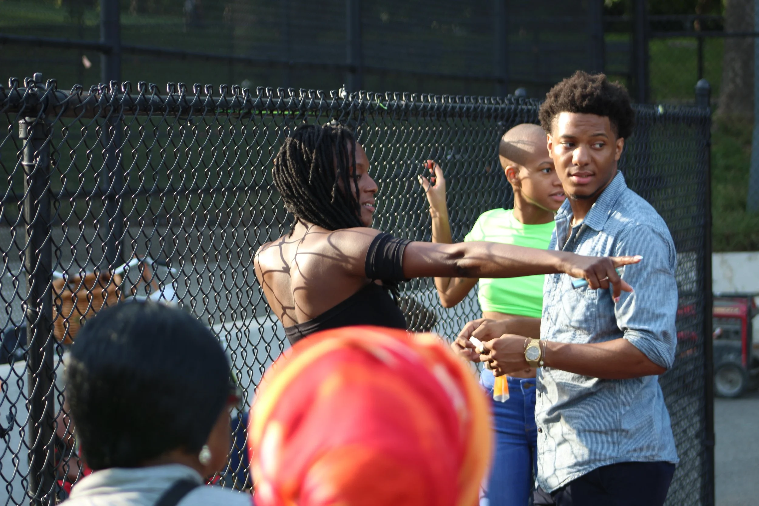 A woman with braided hair is talking to two young boys by a chain-link fence, with a girl in a neon green shirt in the background, outdoors in a park or sports area.