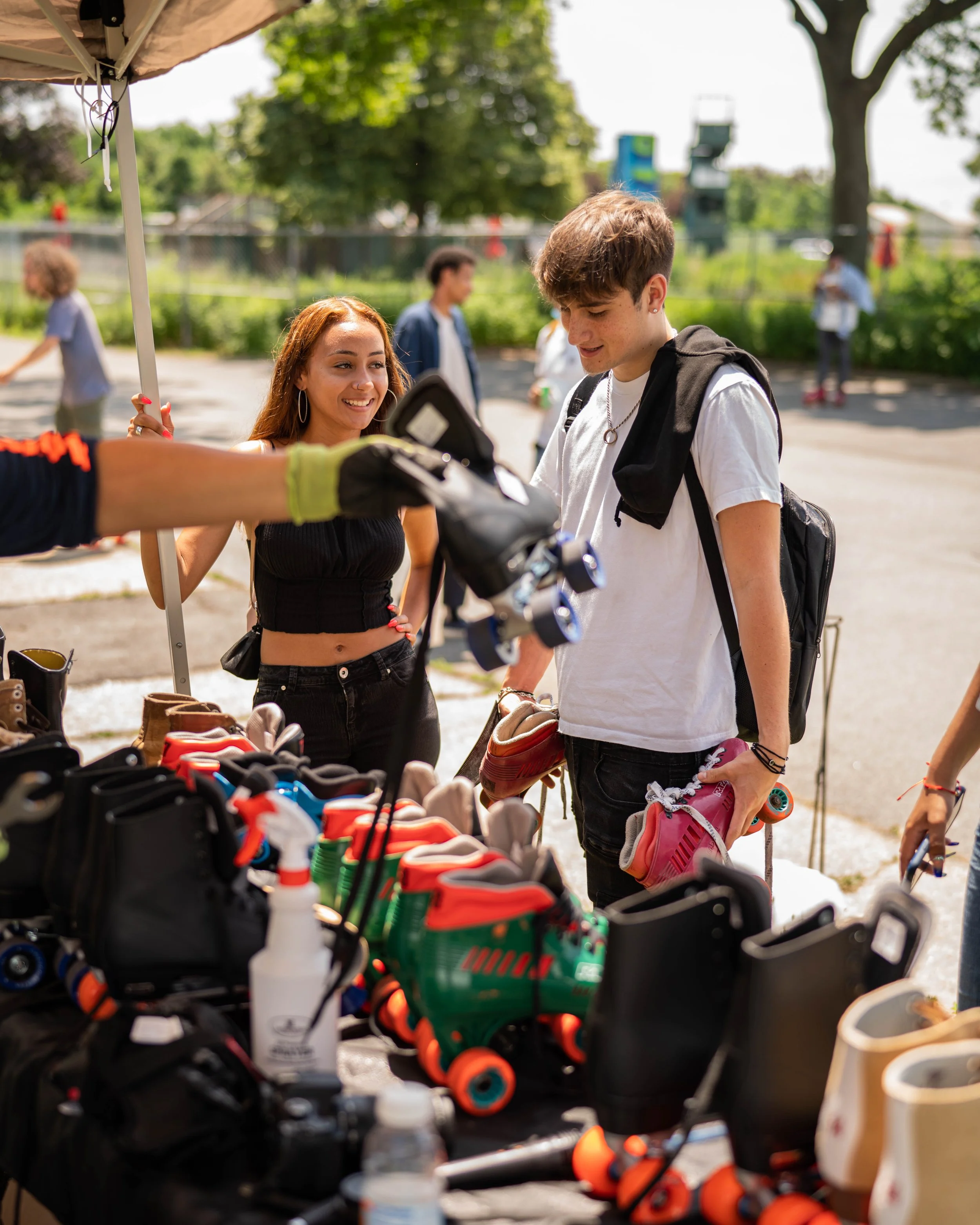 Two young people shopping for roller skates at an outdoor market stall, with shelves filled with various roller skates in the foreground.