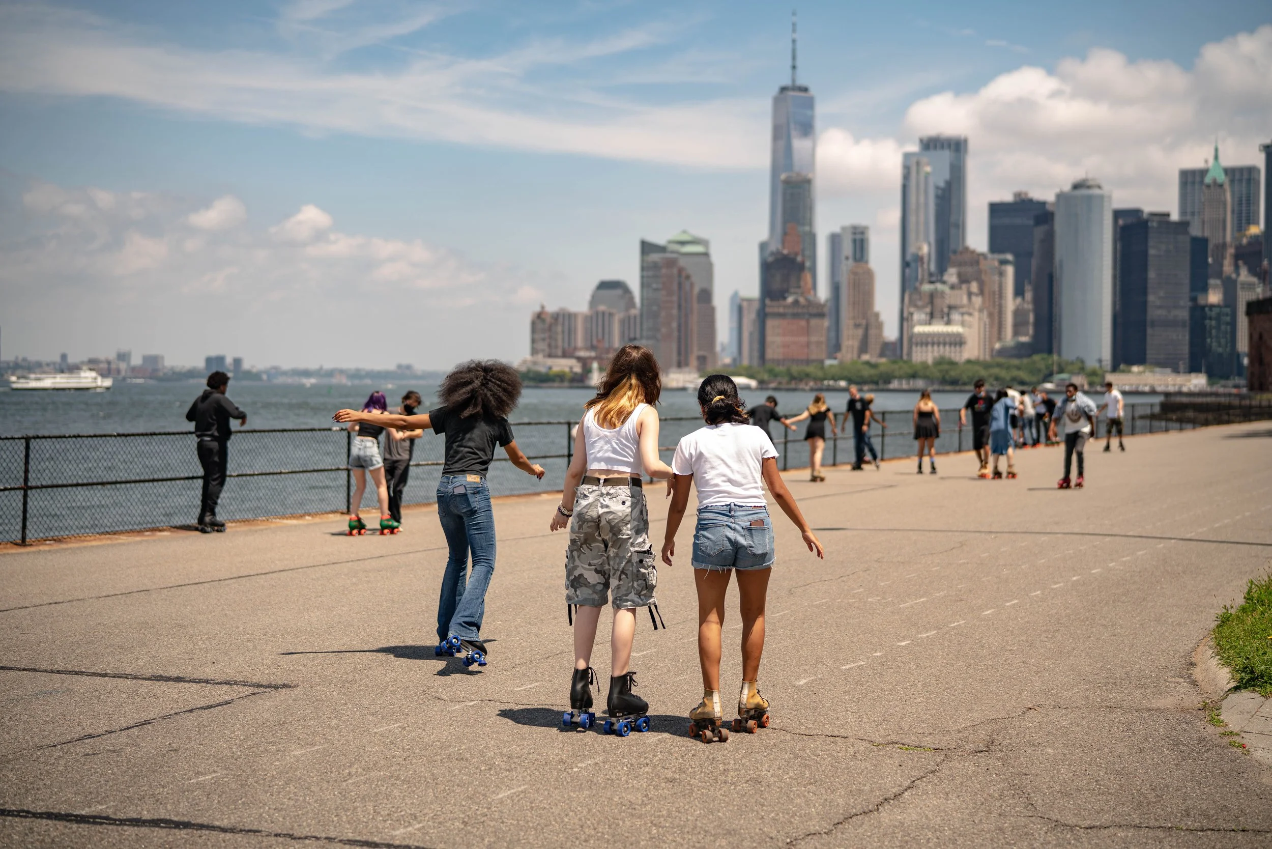 People roller skating along a waterfront promenade with a city skyline, including the One World Trade Center, in the background.