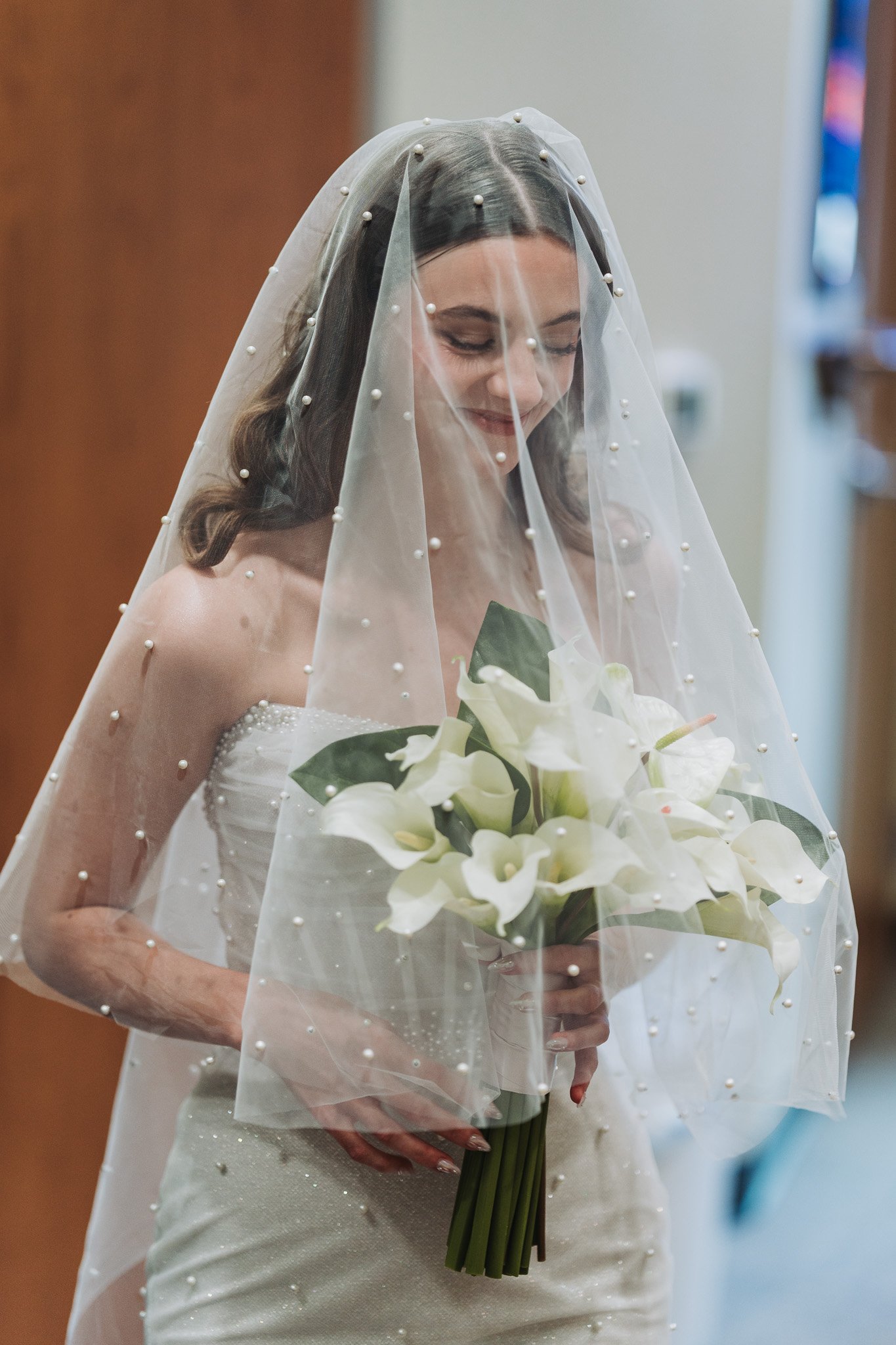 A bride with dark hair in loose waves, wearing a strapless wedding dress adorned with small pearls, looking down and smiling gently while holding a bouquet of white calla lilies.