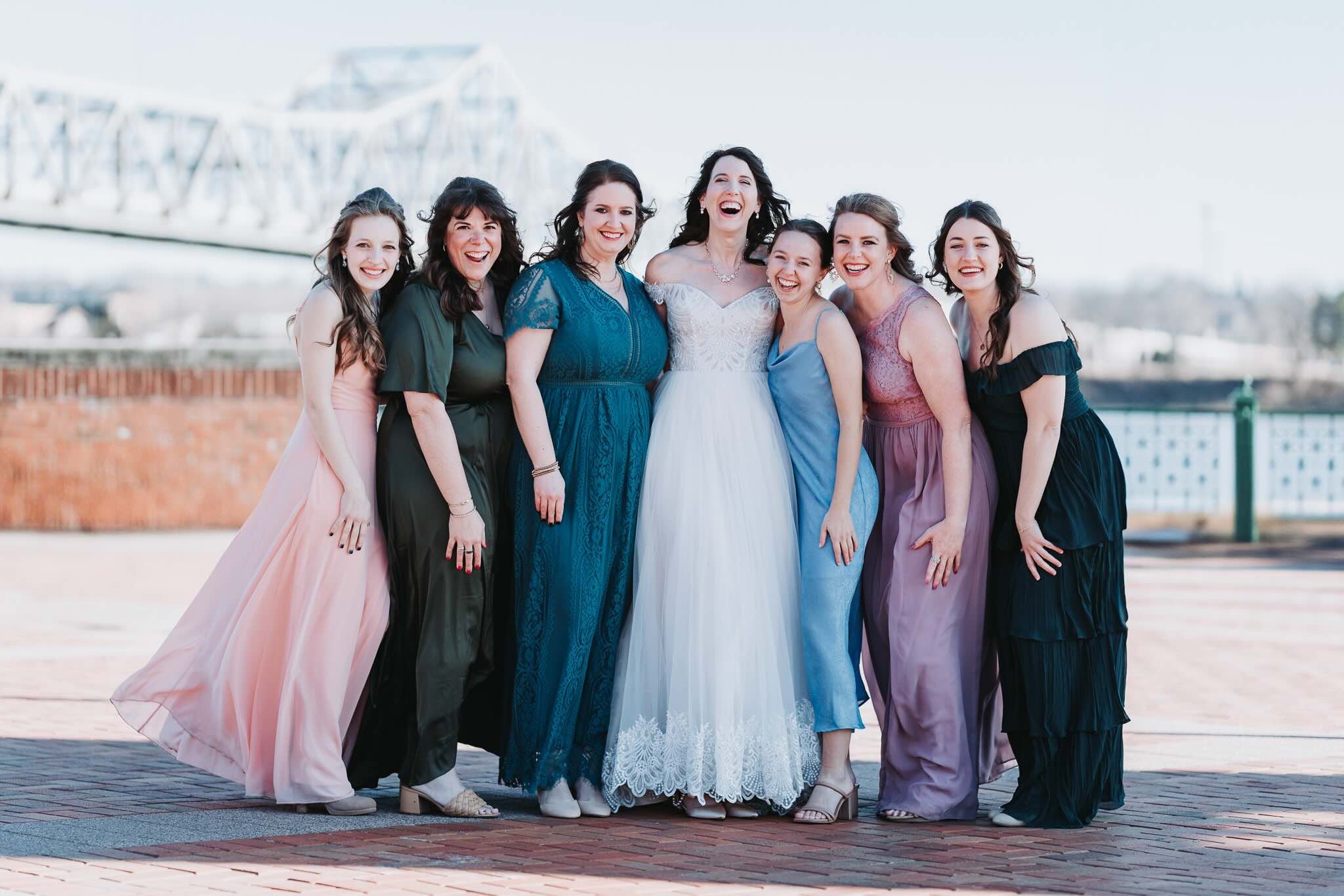 Group of women dressed in colorful dresses, smiling and laughing outdoors, with a bridge in the background.