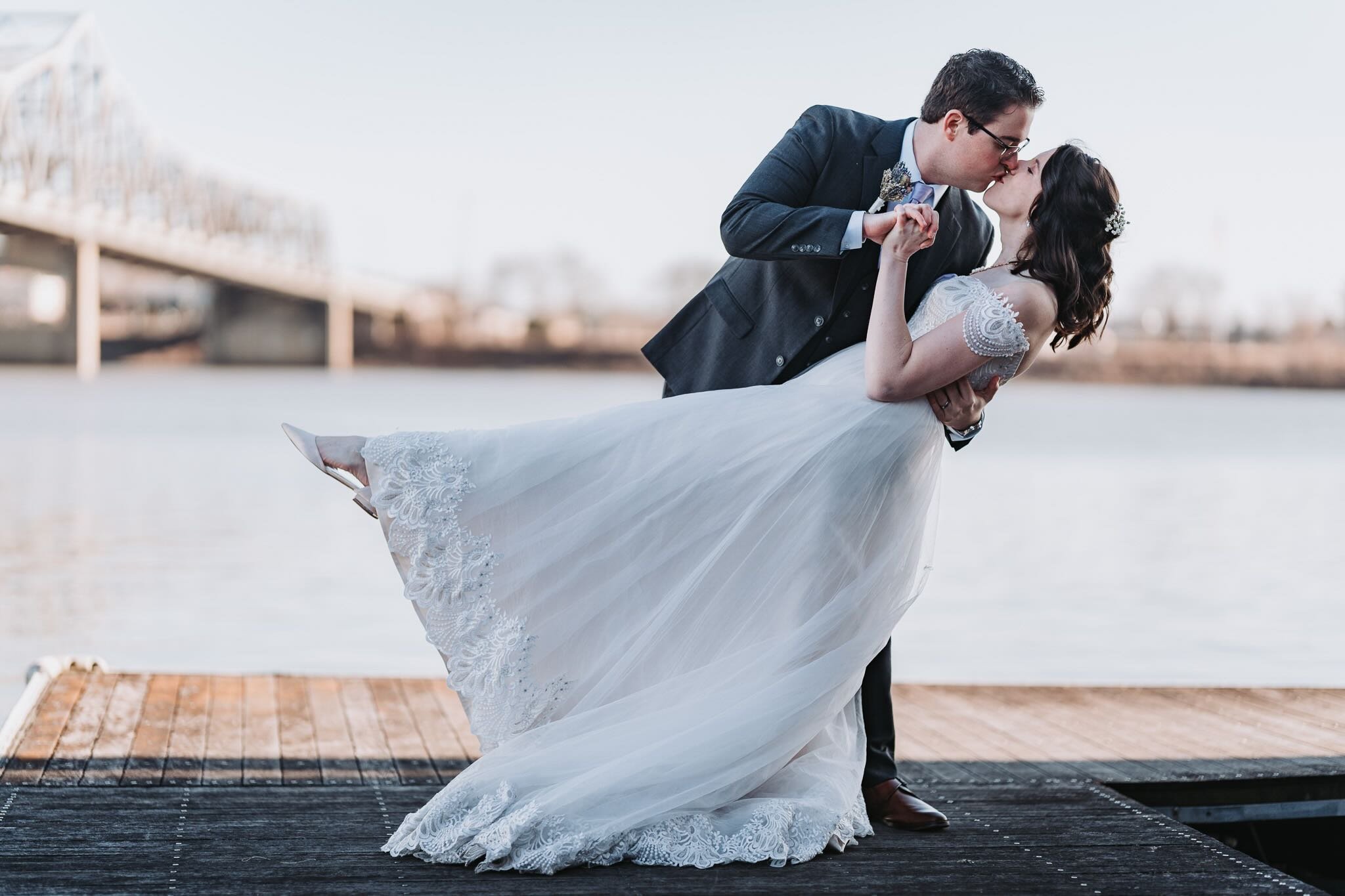 A bride and groom share a kiss near a body of water, with the groom dipping the bride. The bride wears a white lace wedding dress and the groom a dark suit. The background features a bridge and a cloudy sky.