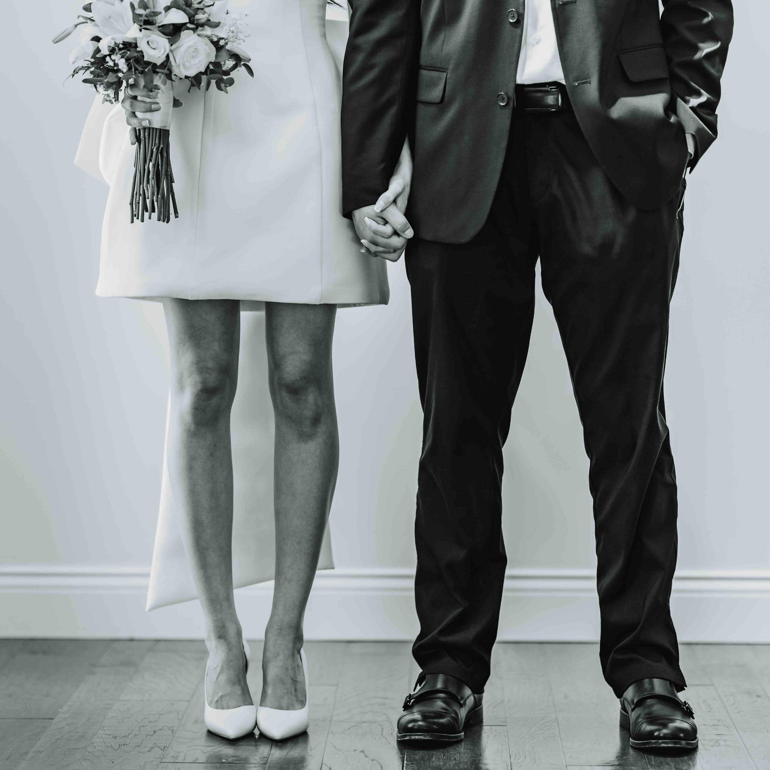 Close-up of a couple holding hands, with the woman wearing a white dress and high heels, and holding a bouquet of flowers, and the man wearing a dark suit. The photo is in black and white.
