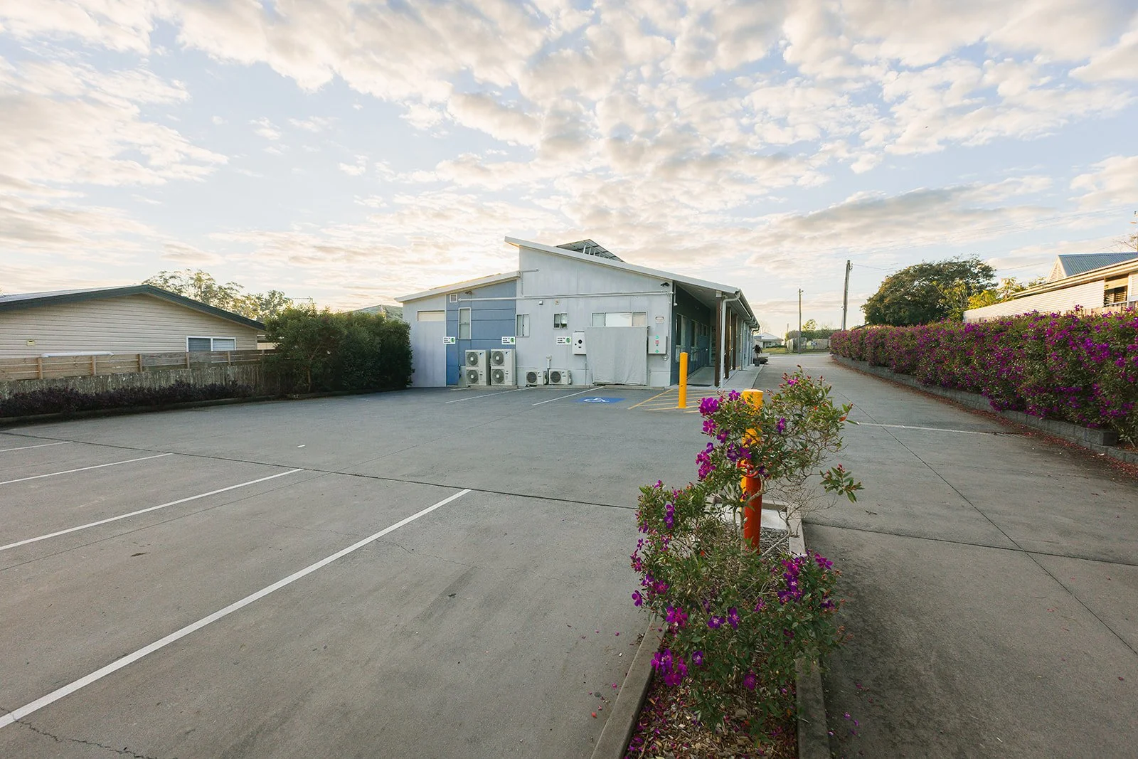 Empty parking lot with a building in the background, flowering bushes along the right side, and a partly cloudy sky overhead.